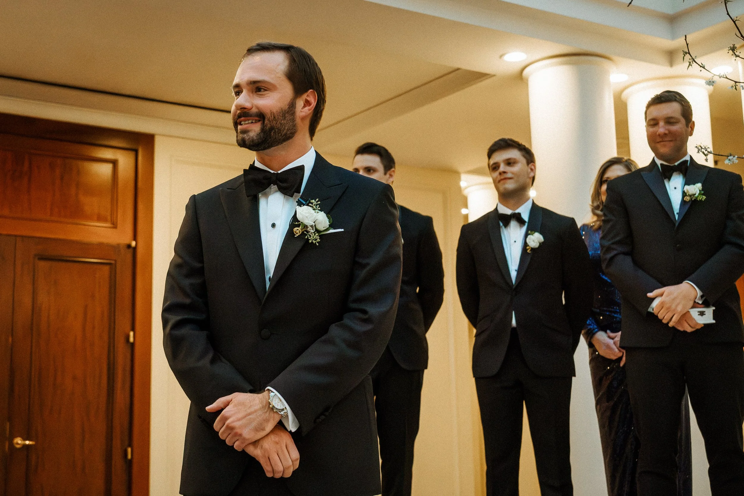 A groom in a tuxedo with a boutonniere is standing with his hands clasped in front of him, smiling, with four other men in tuxedos and a woman in a formal dress standing behind him in an indoor setting with warm lighting and a wooden door.