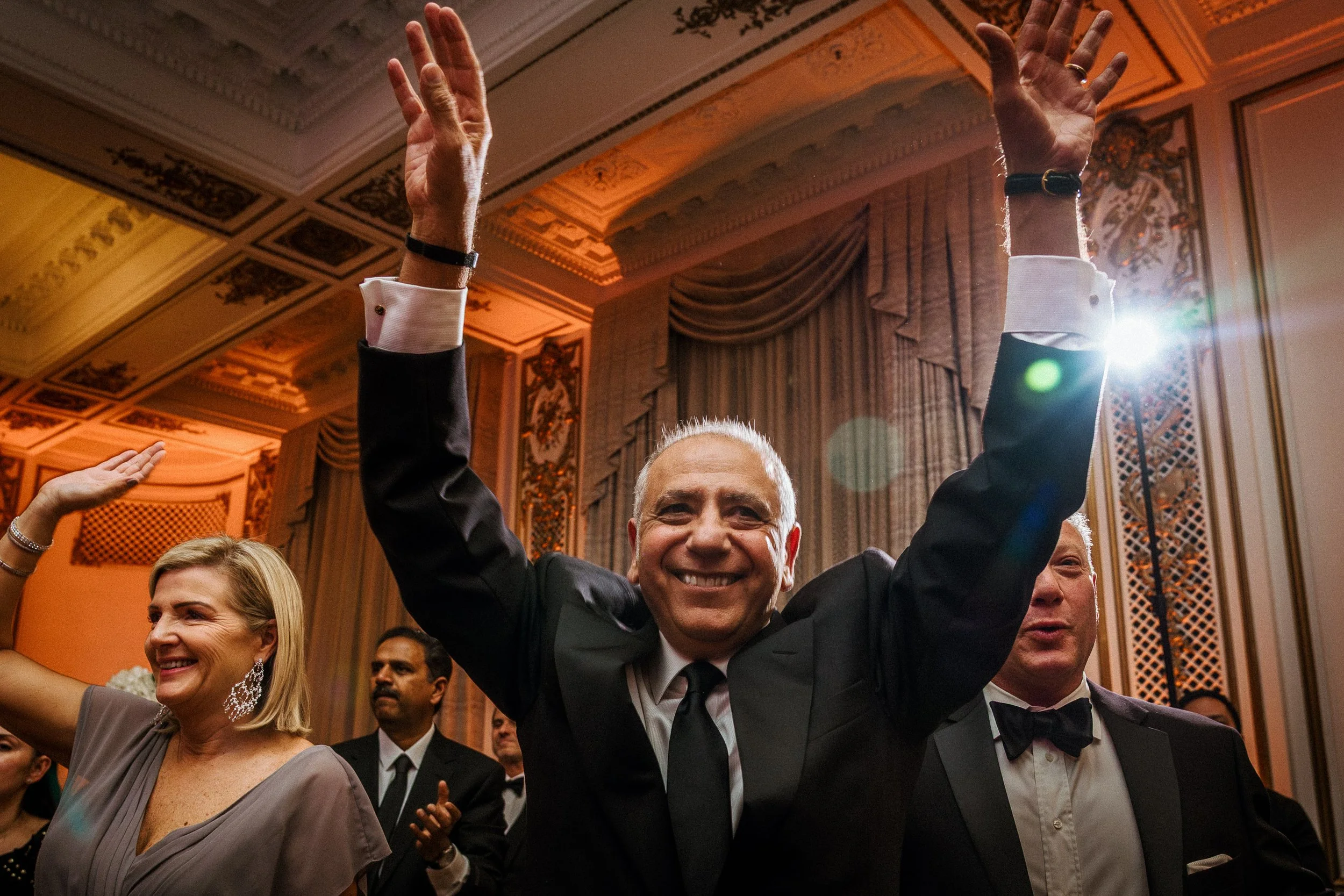 Smiling man in a tuxedo raising his hands at a formal event in a decorated ballroom.