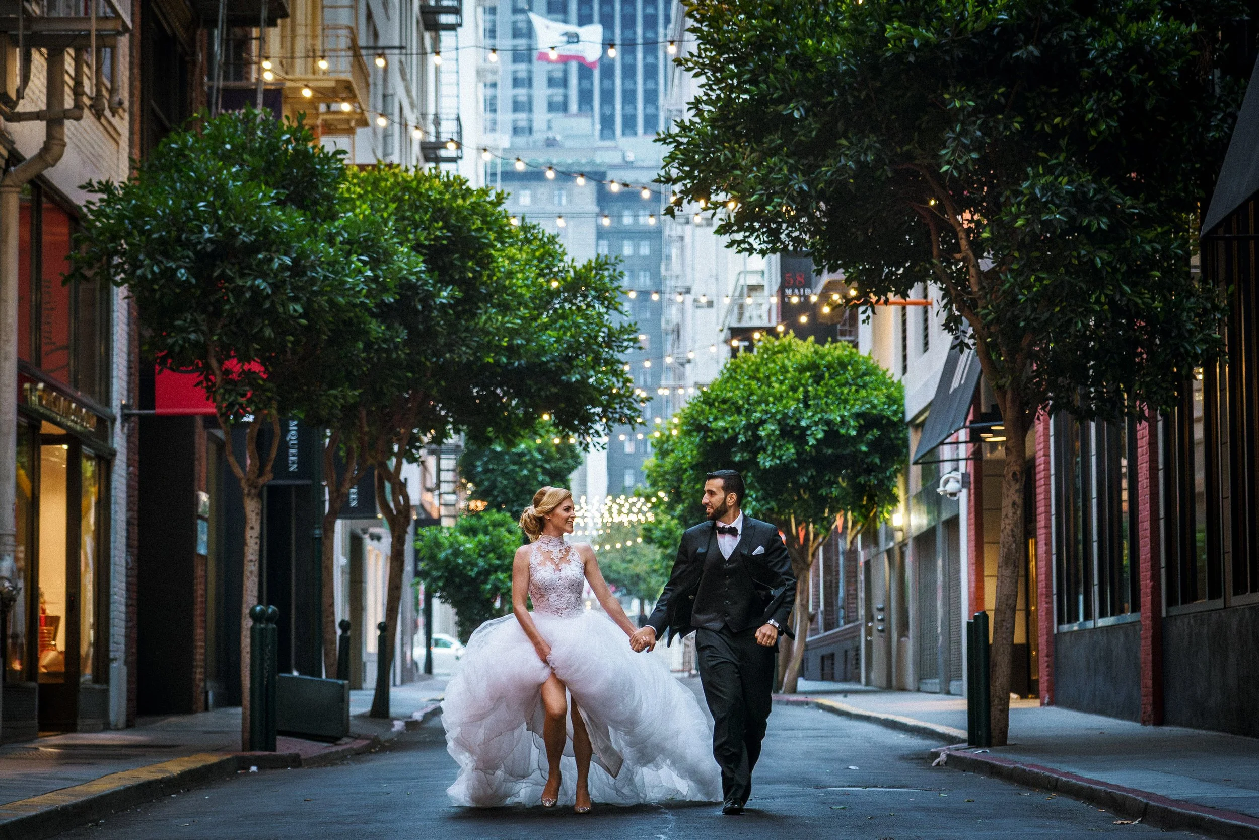 A newlywed couple holding hands and walking hand in hand down a city street at dusk. The bride is wearing a white wedding dress with a lace top and a full skirt. The groom is in a black tuxedo. They are in San Francisco.