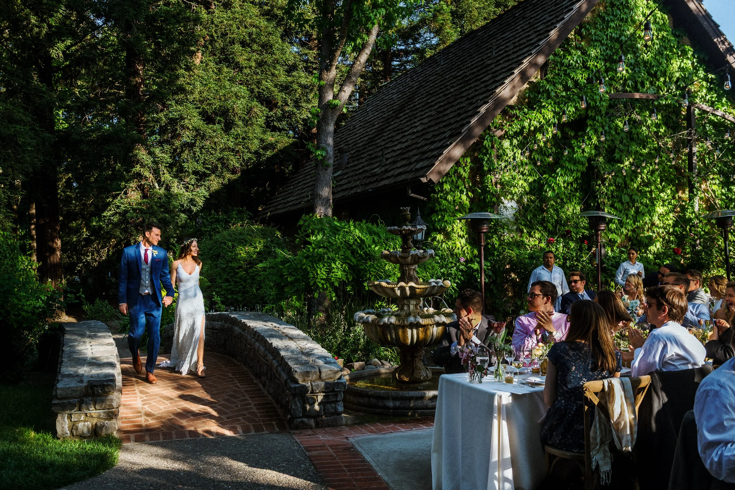 A wedding reception outdoors with guests seated at tables, a fountain, and a bride and groom walking on a brick path in a lush garden setting.