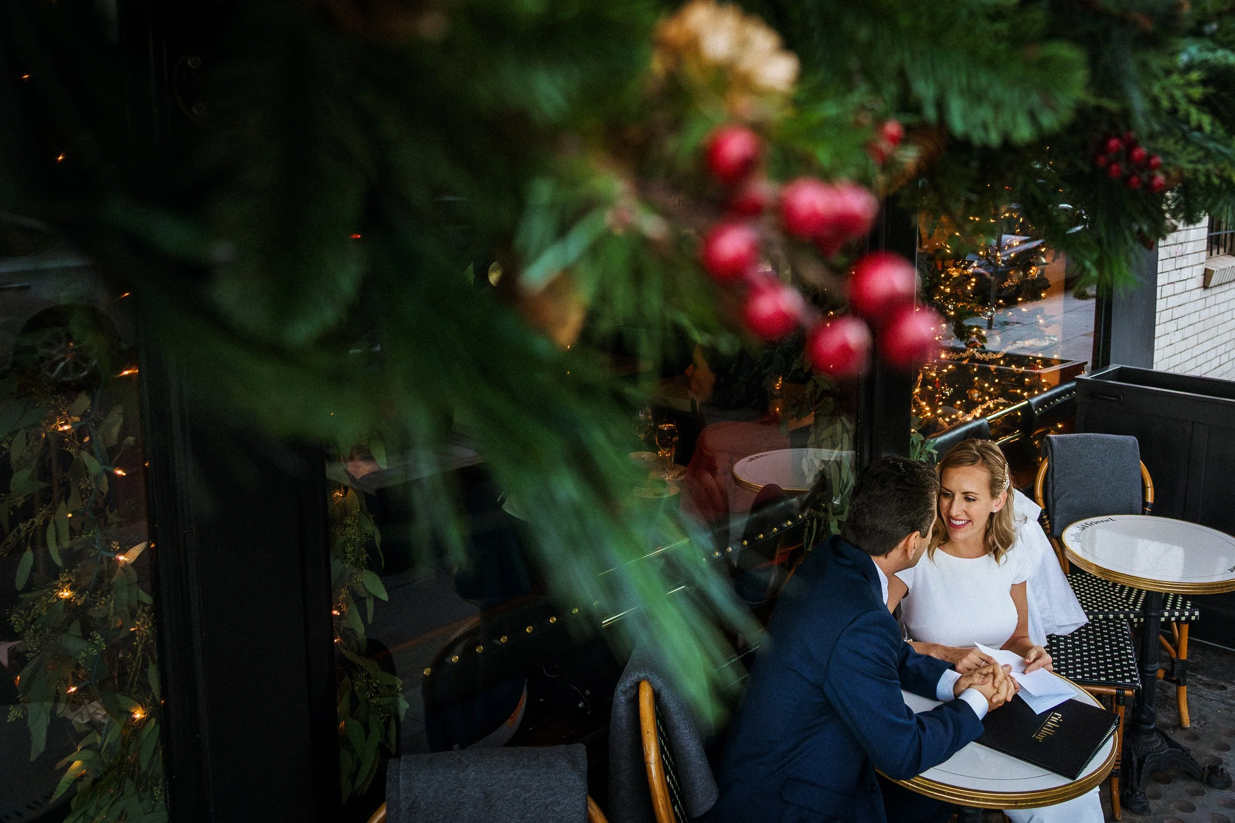 A couple talking and holding hands at a restaurant table decorated for Christmas with greenery and red berries, seen through the leaves and berries in the foreground.
