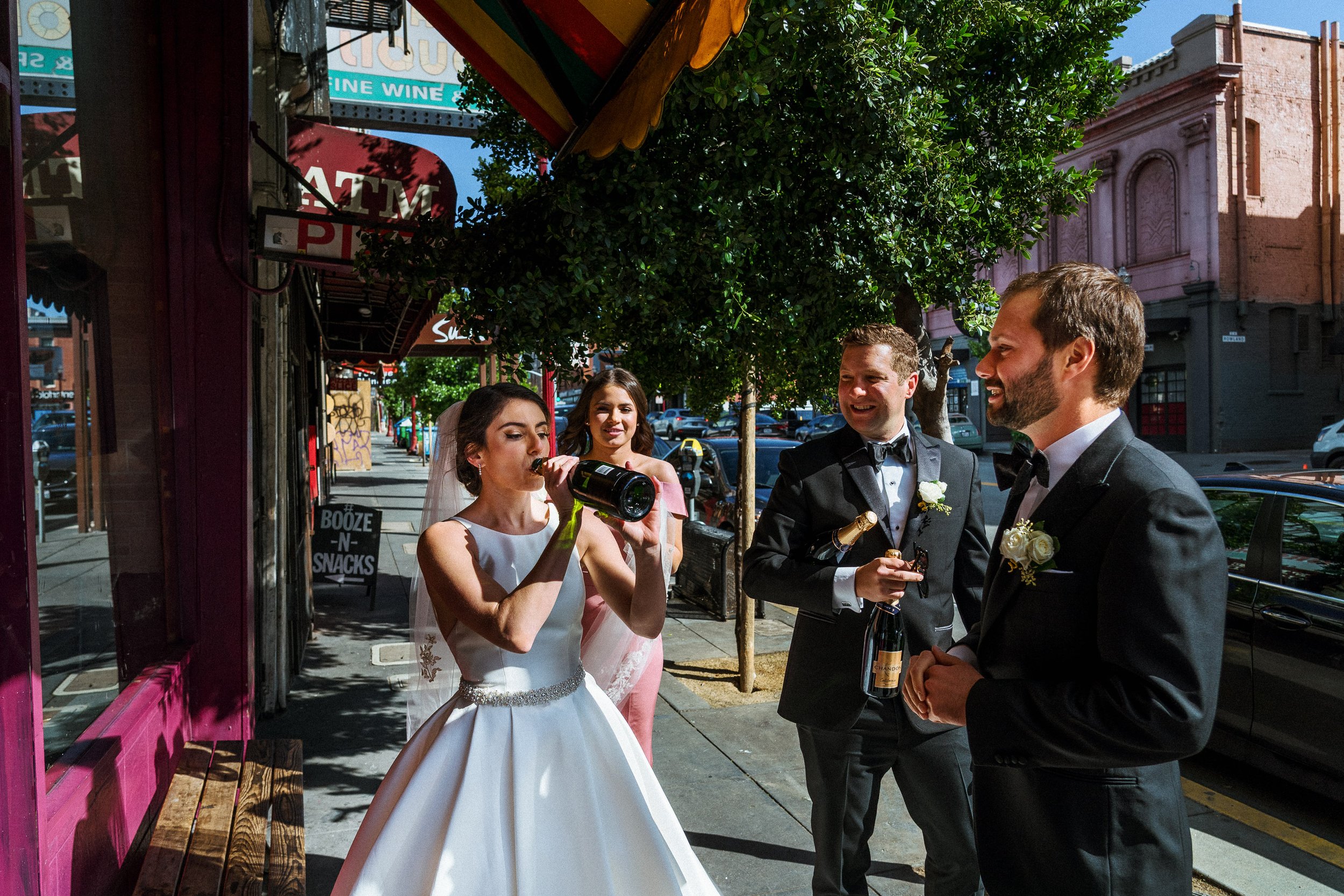 A bride in a white wedding dress drinks from a bottle while three groomsmen in tuxedos stand nearby smiling on a sunny city sidewalk.