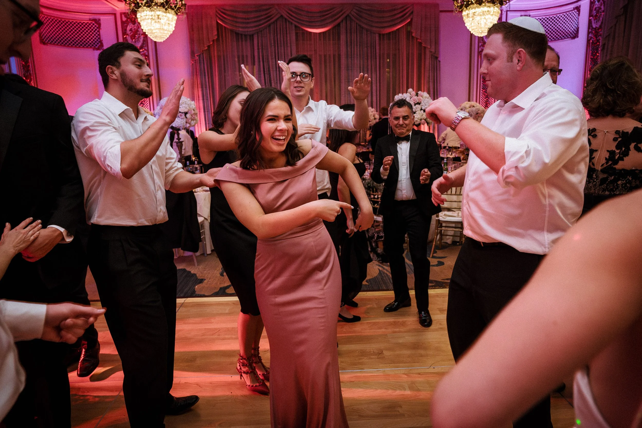 People dancing and having fun at a wedding reception in an elegant, decorated ballroom with draped curtains and chandeliers.
