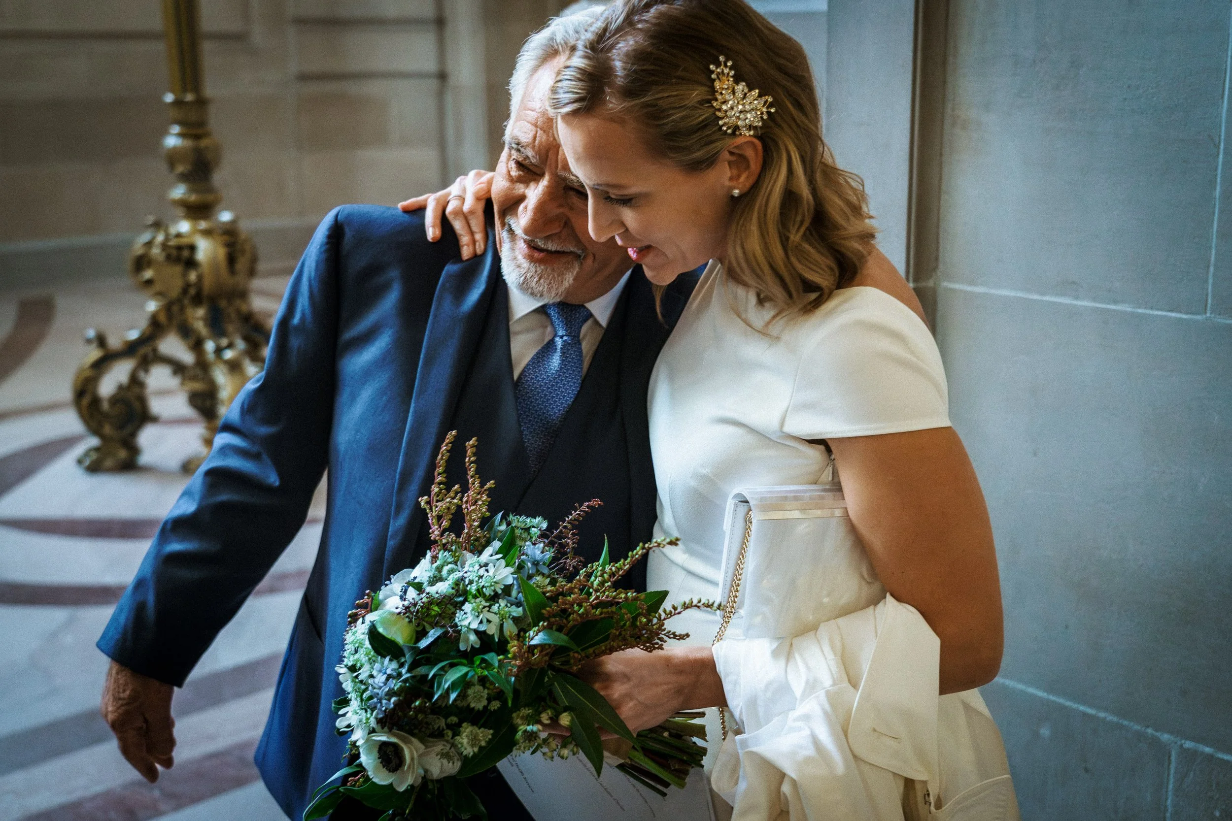 A young woman in a white dress embraces an older man in a dark suit, both smiling warmly, with the woman holding a bouquet of flowers.
