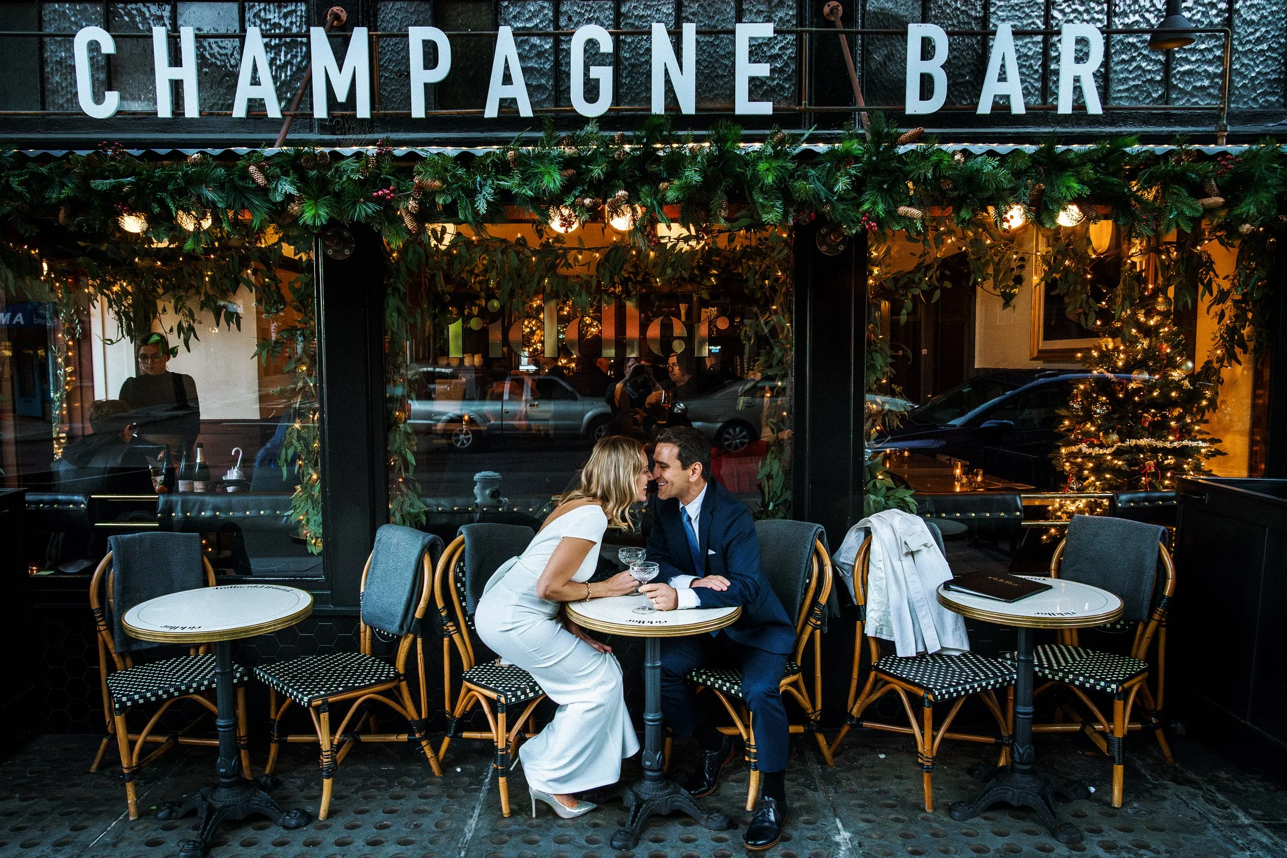 A couple sitting at a small round table outside a champagne bar, leaning in toward each other, smiling, with glasses of champagne, surrounded by festive holiday decorations, including a small Christmas tree and greenery with string lights.