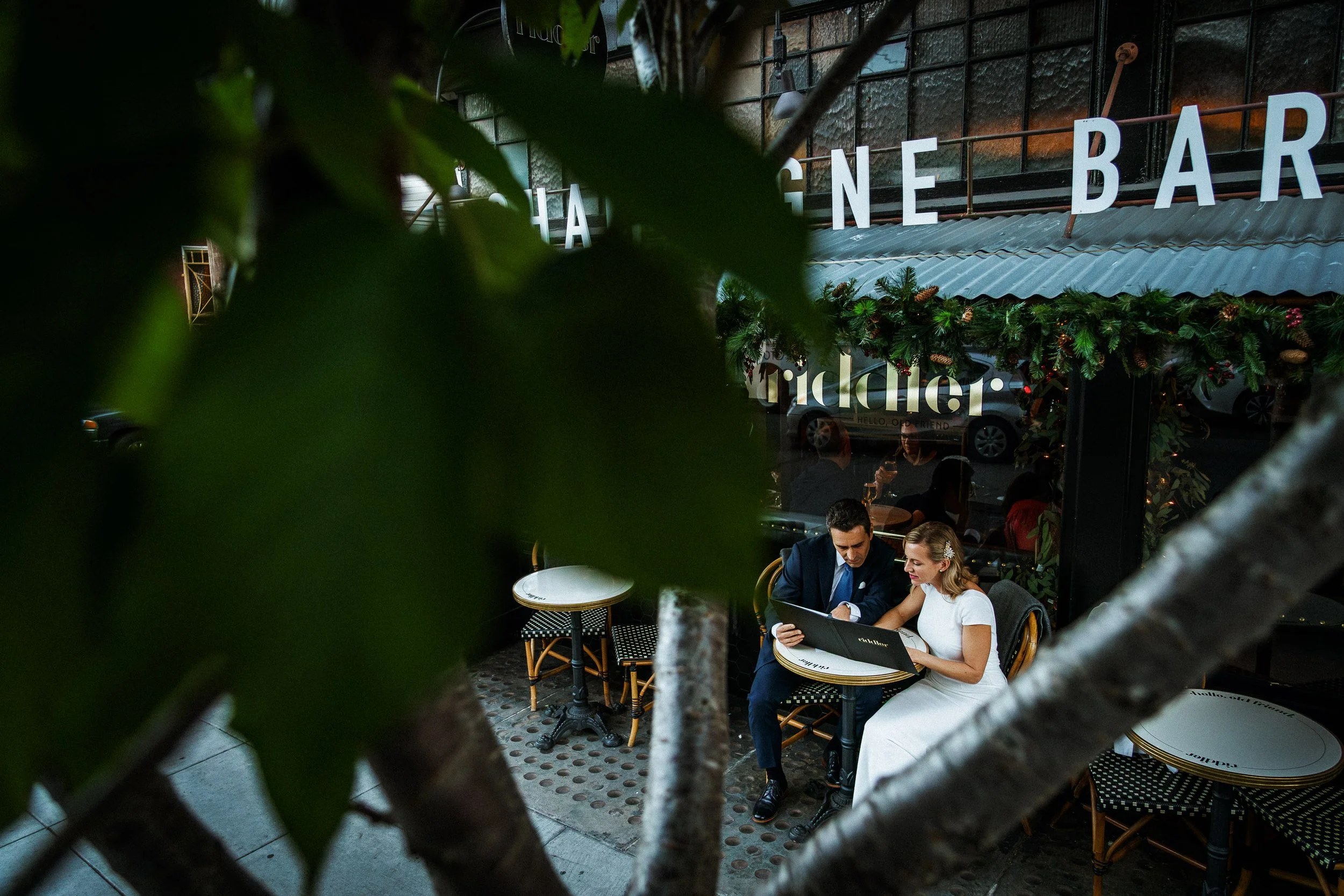 Couple in formal attire sitting at an outdoor table in a restaurant, looking at a menu, with decorative greenery and lights above, seen through tree branches and leaves.