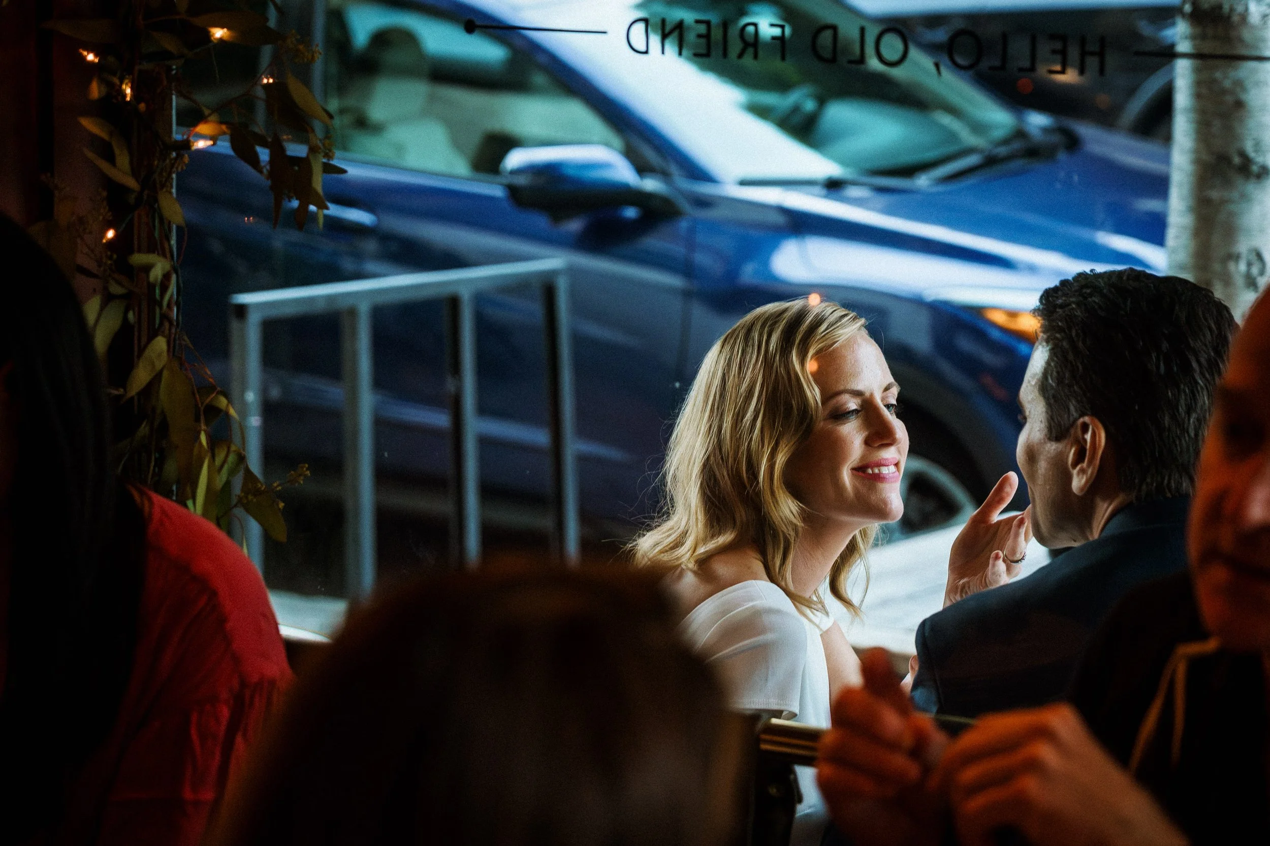 A woman and a man are sitting together at a restaurant with a window view of parked cars outside. The woman is smiling with her eyes closed, and the man is looking at her, touching her face gently.