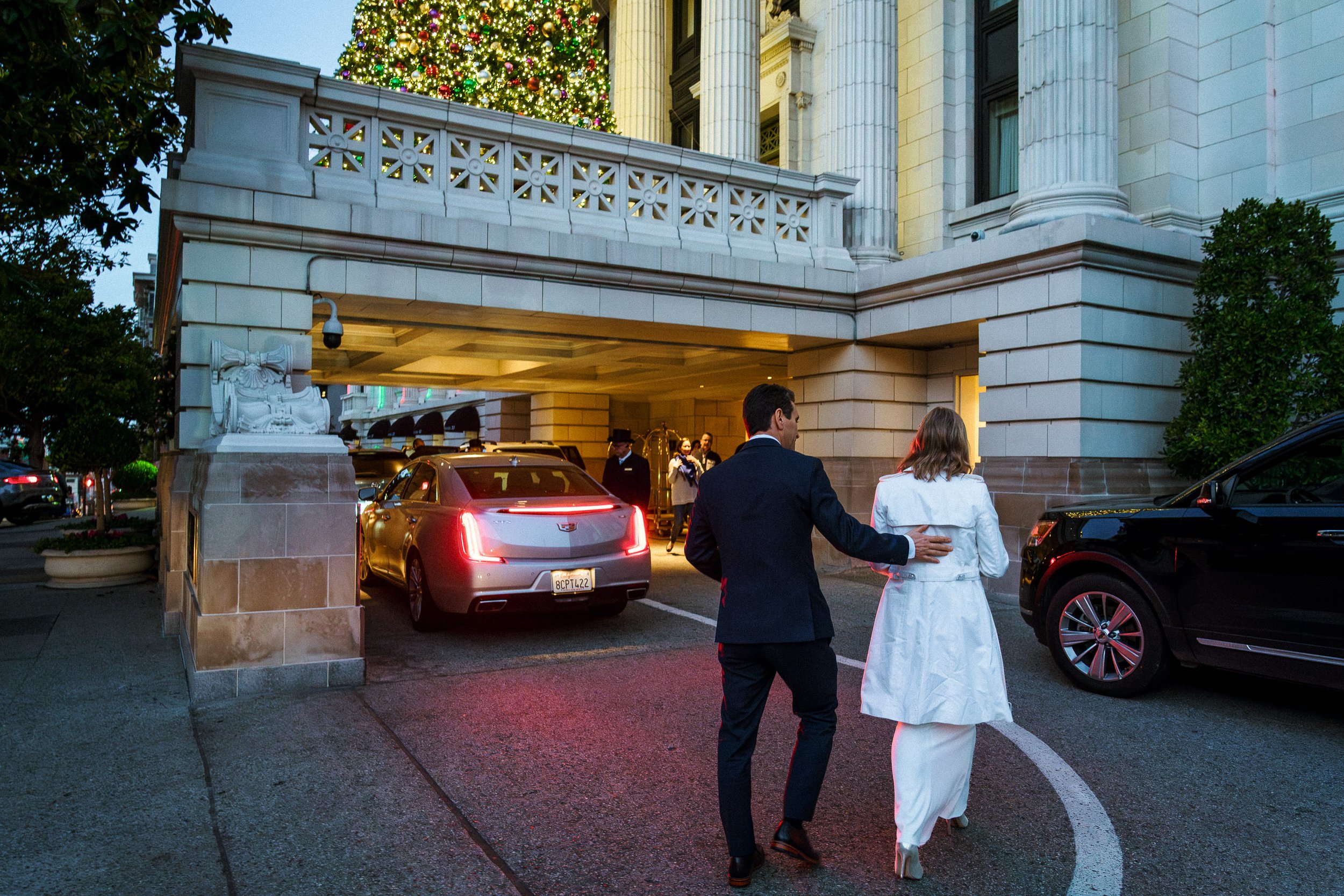 A man and woman walking towards a grand building with a Christmas tree visible on the balcony. The man has his hand on the woman's back, and they are dressed in formal attire.