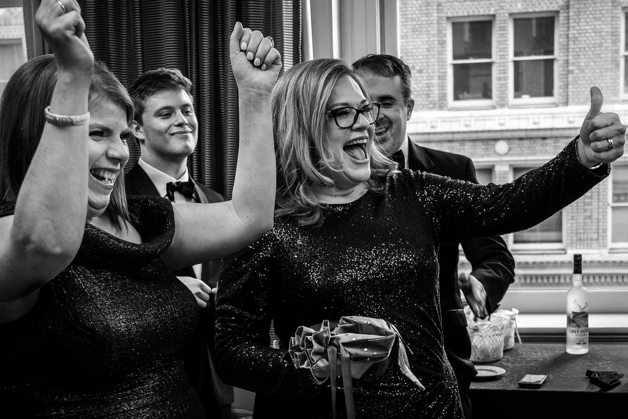 Group of people celebrating, smiling, and cheering at a festive party, some in formal attire, near a window with a cityscape background.