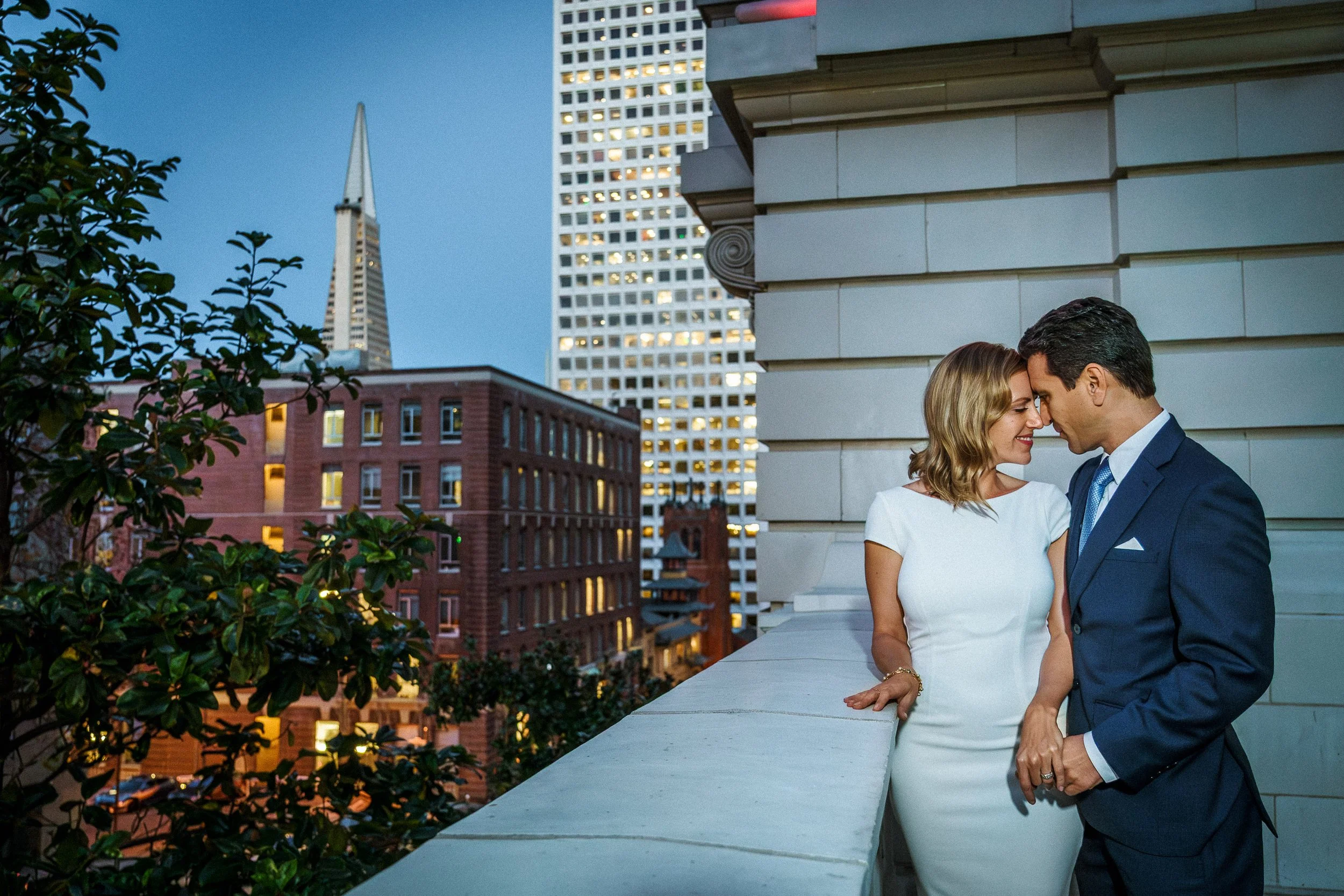 A couple dressed in formal attire on a balcony, with their foreheads touching in an intimate pose, city skyline with tall buildings and the Transamerica Pyramid in San Francisco in the background during evening.
