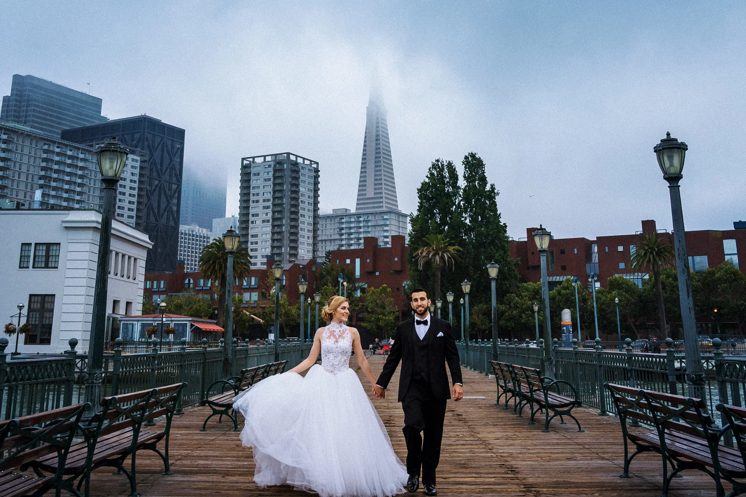 A bride and groom walking hand in hand on a wooden pier with city buildings and the Transamerica Pyramid in the foggy background.