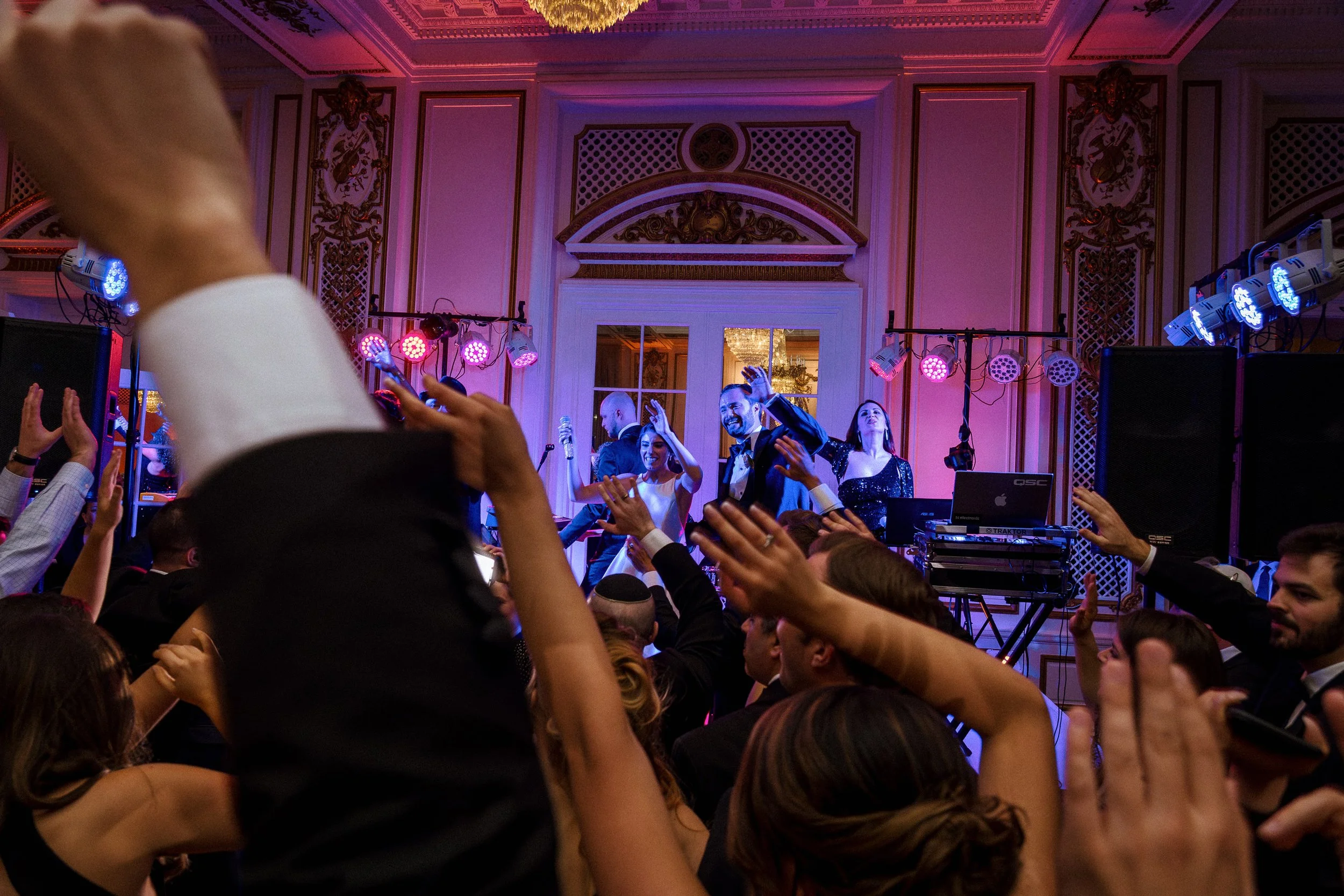 People dancing and enjoying a lively celebration in an ornate ballroom with colorful lighting and a DJ setup.
