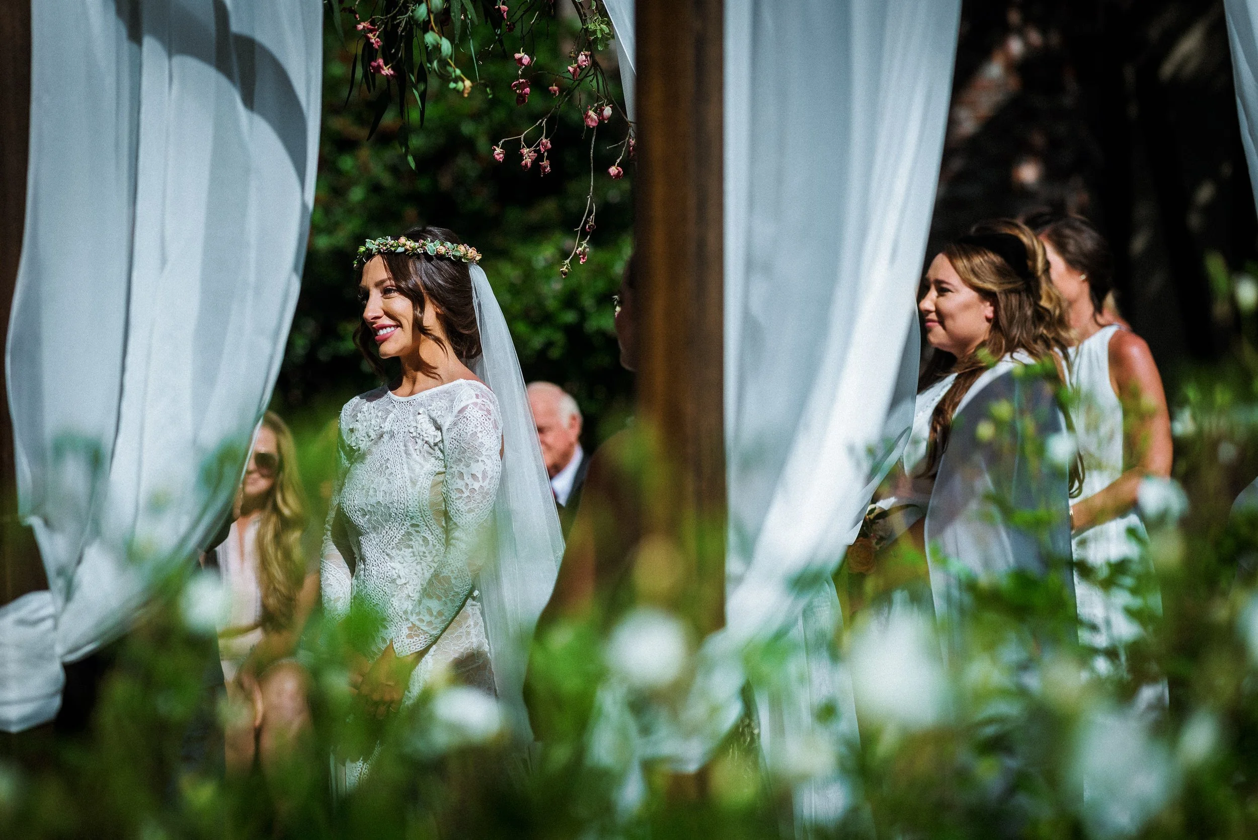 Bride in a white lace wedding dress smiling during an outdoor wedding ceremony, surrounded by greenery and white drapes.