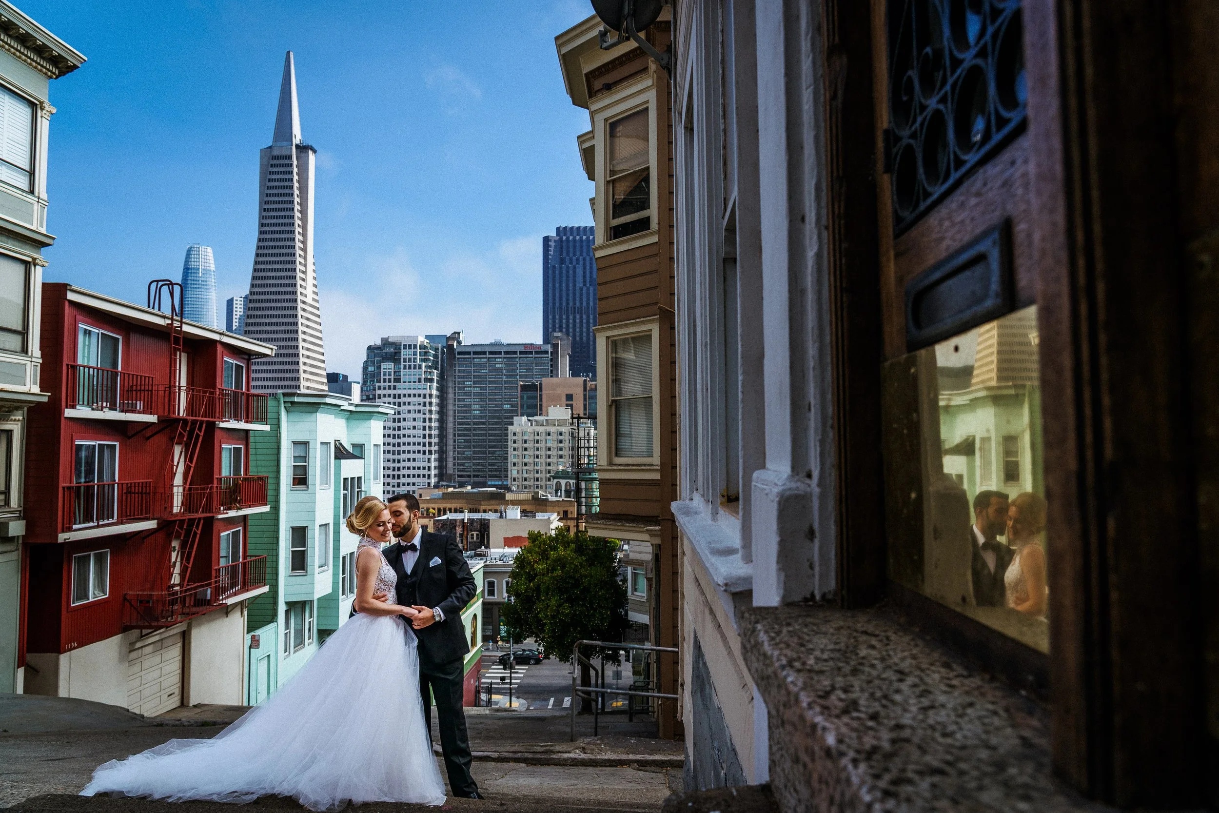 A bride and groom standing on a city street in San Francisco, with colorful houses in the foreground and tall skyscrapers in the background, reflected in a window on the right.