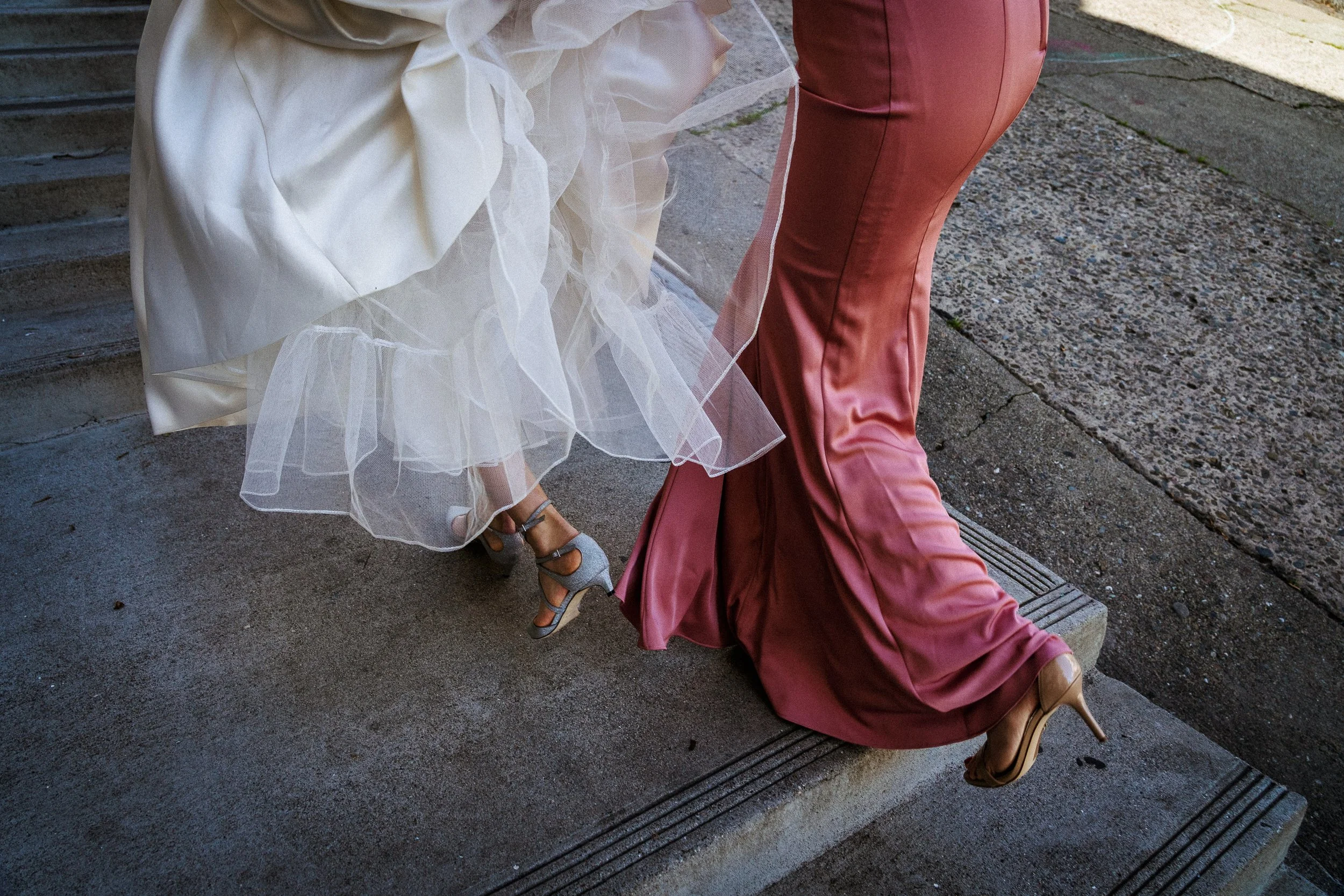 A bride in a white wedding dress walking down the steps, accompanied by a bridesmaid in a satin pink gown, both wearing high heels.