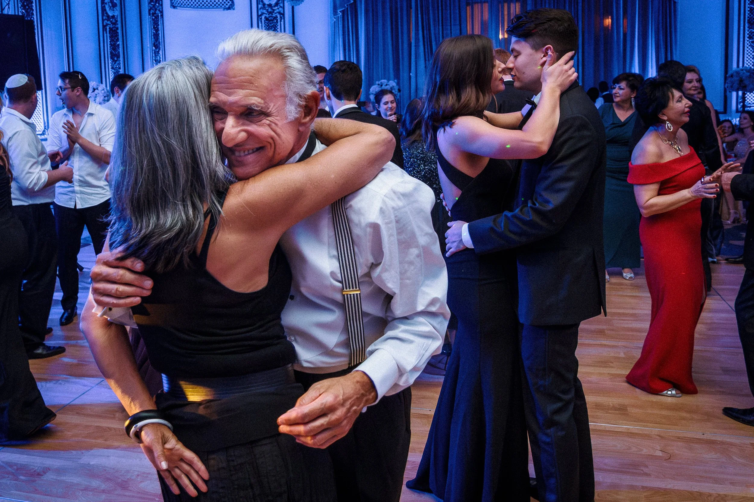People dancing and hugging at a formal event, with couples embracing closely on a wooden dance floor, in an elegant decorated room with blue lighting.