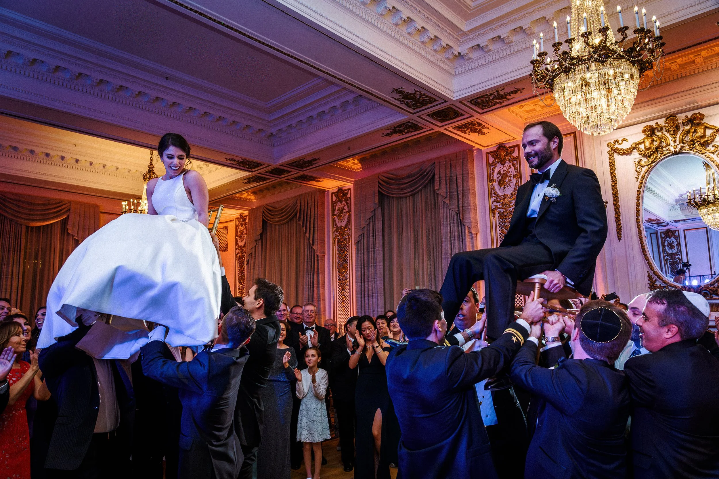 Wedding reception with a bride and groom sitting on chairs carried by guests during the hora dance in an elegantly decorated ballroom.