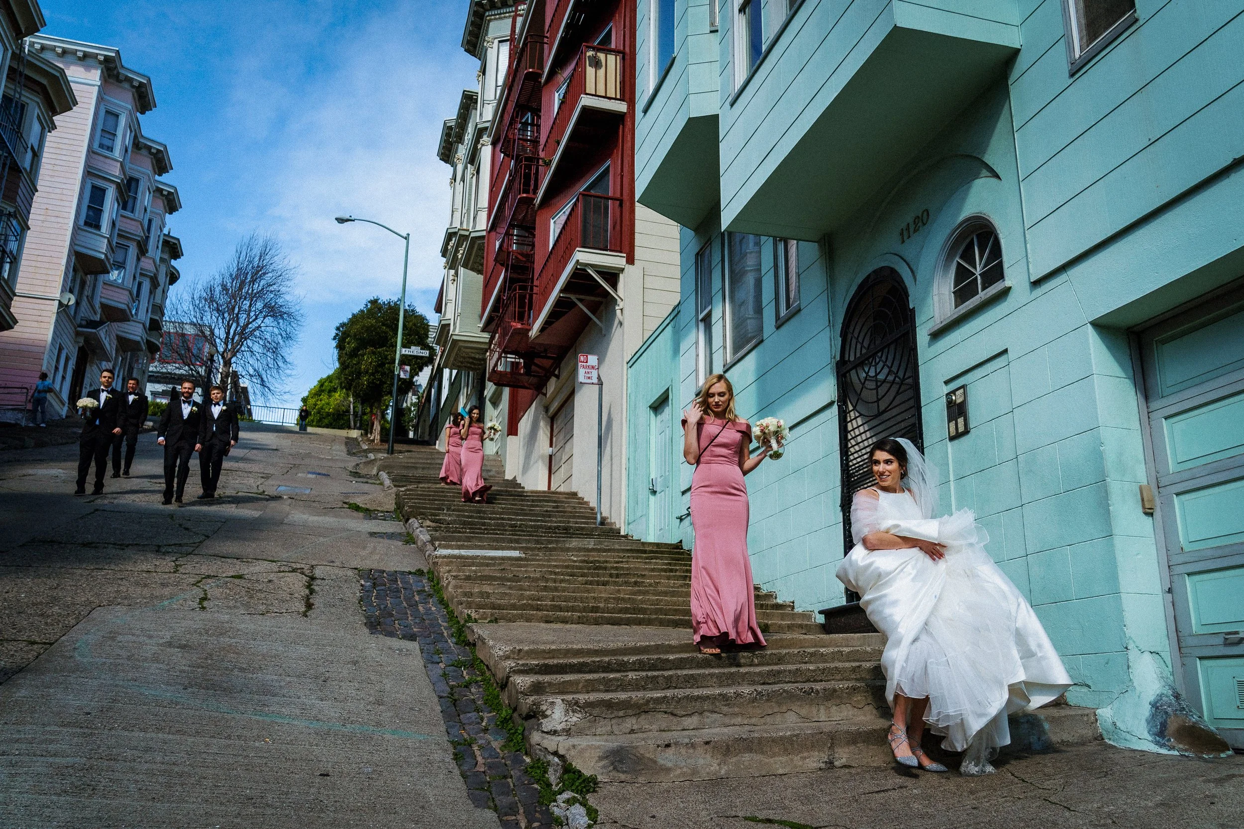 A bride in a white wedding dress and veil sitting on steps outside a teal-colored building, with bridesmaids in pink dresses holding bouquets beside her. A group of groomsmen in suits walks down the street in the background.