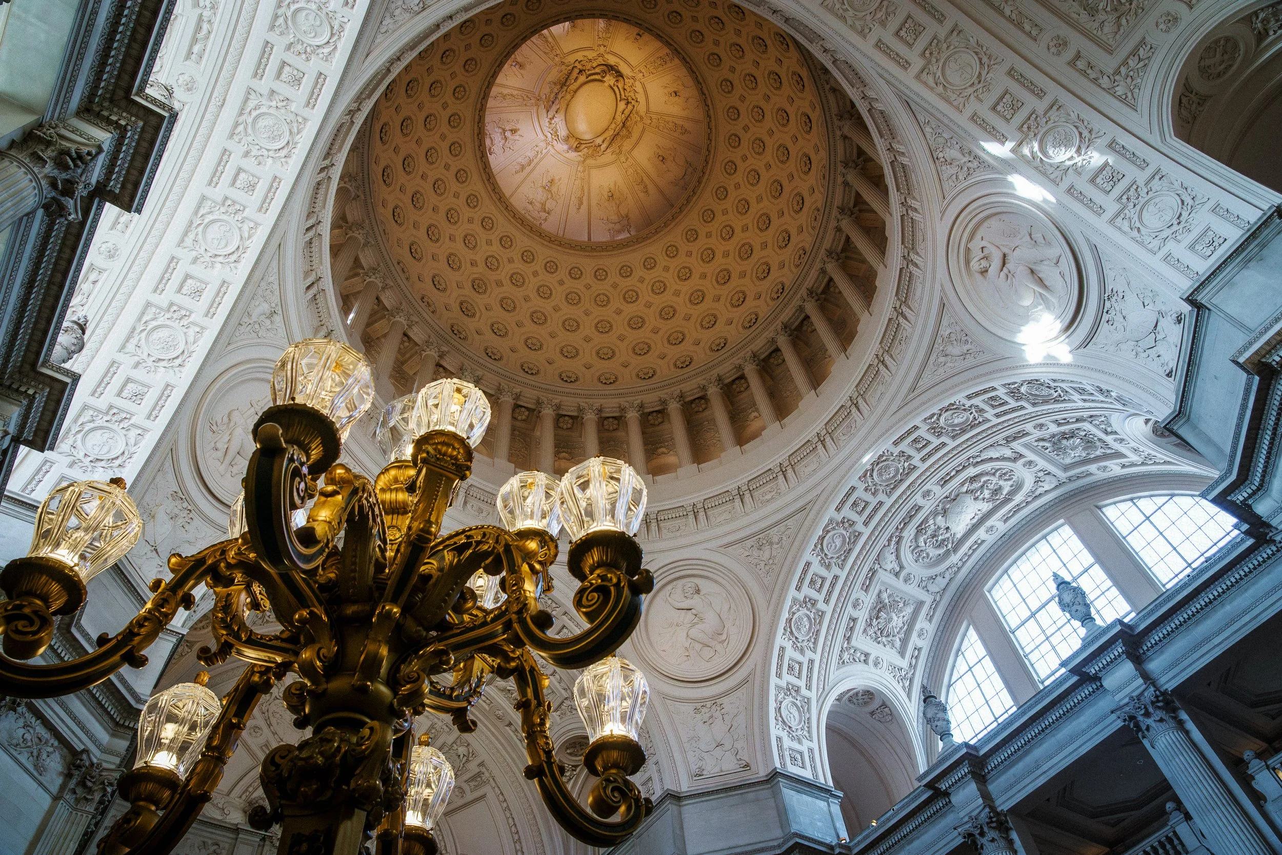 Interior view of a grand, ornate building with a high domed ceiling, intricate white plaster moldings, large arched windows, and a gold chandelier hanging from the ceiling. San Francisco City Hall.