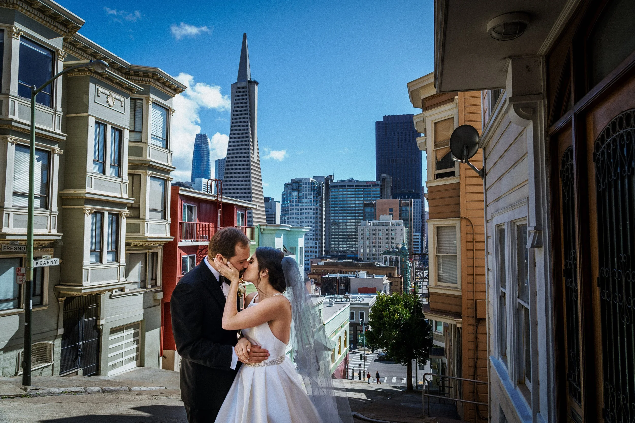 A bride and groom kissing on a city street in front of colorful houses with San Francisco's skyline and skyscrapers in the background.