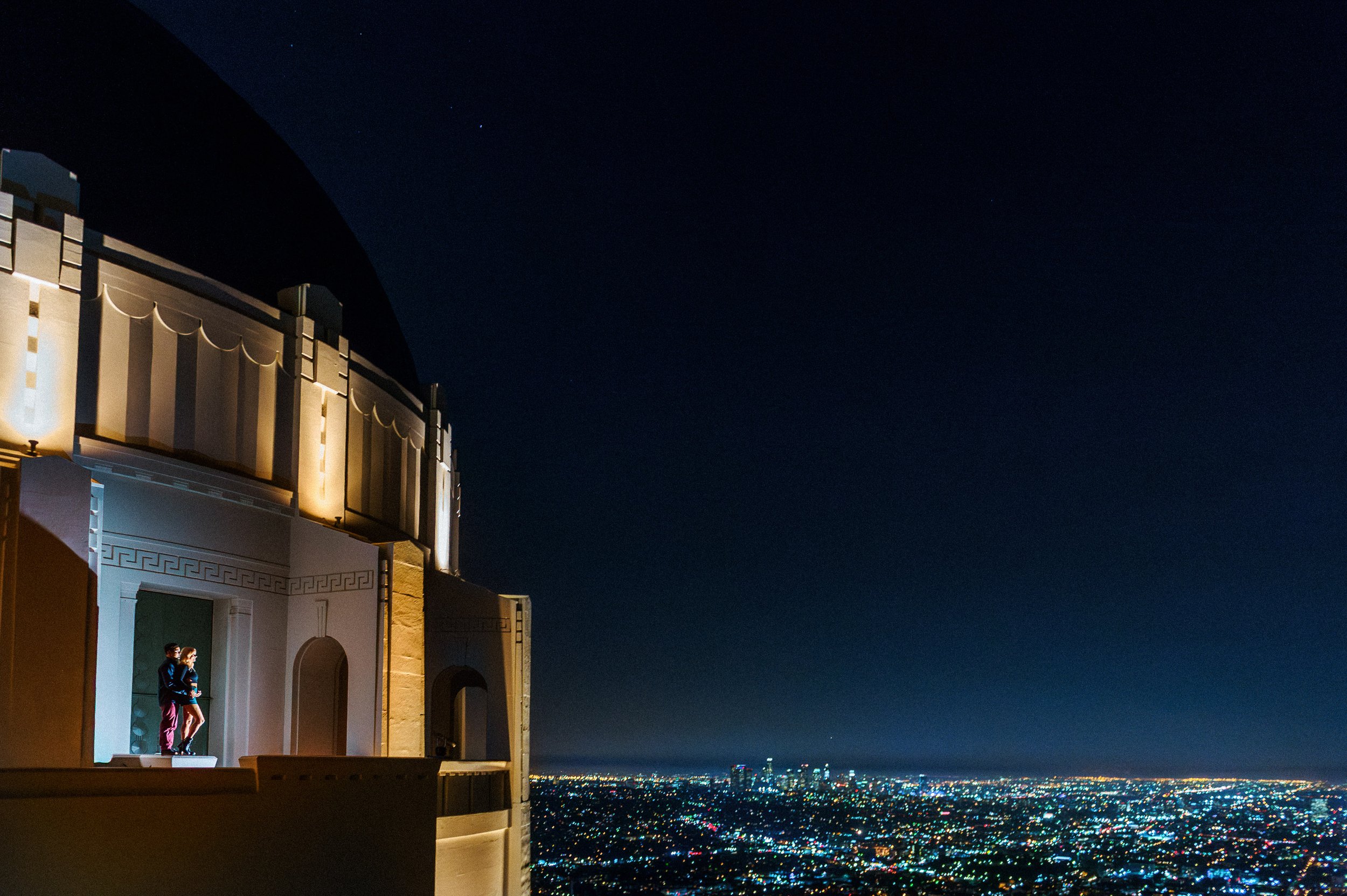 Nighttime engagement session at the Griffith Observatory in Los Angeles, showing a couple embracing on a balcony with the city lights ahead. Engagement photo.