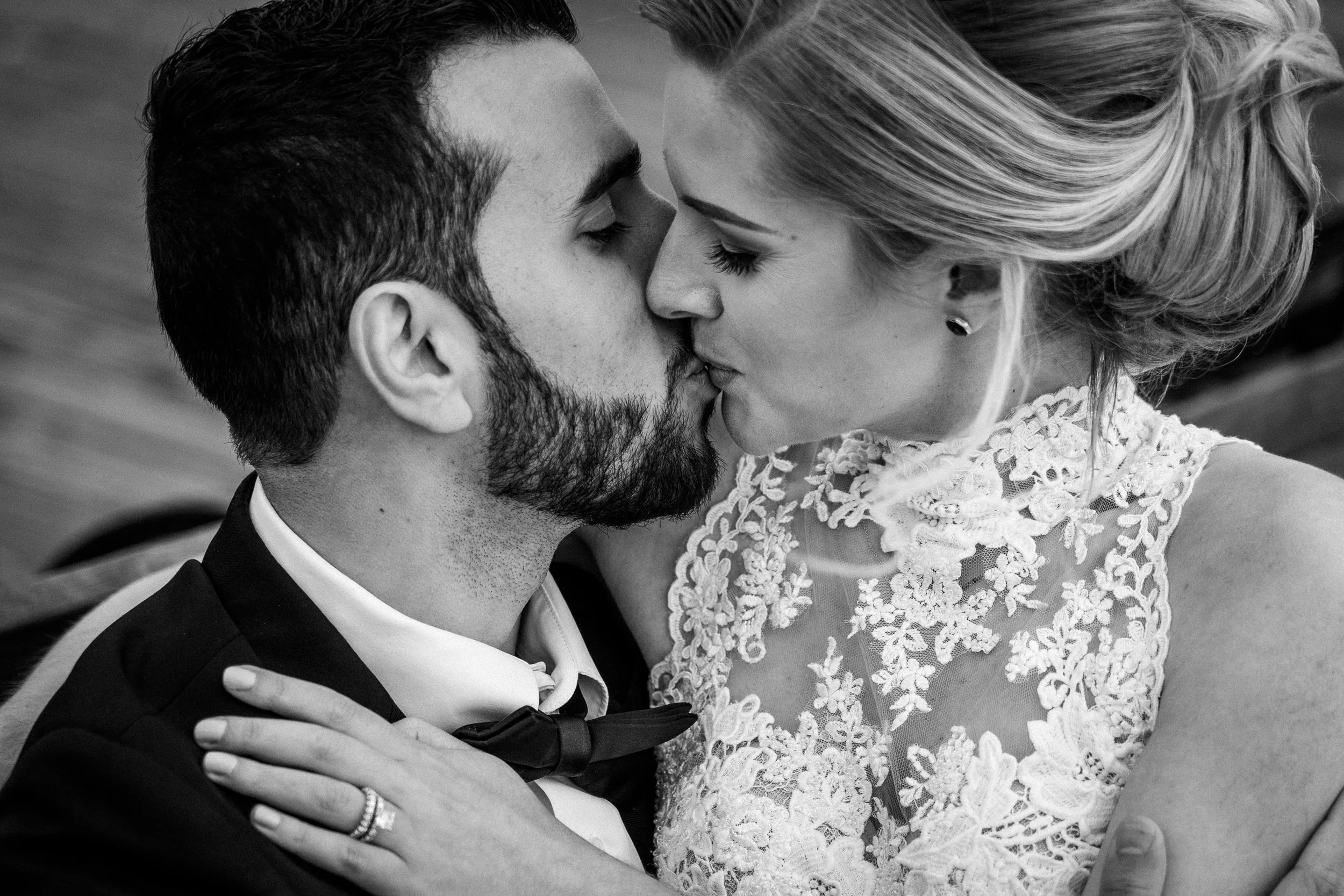 A black and white close-up photo of a couple kissing, with the woman wearing a lace dress and the man in a tuxedo with a bow tie.