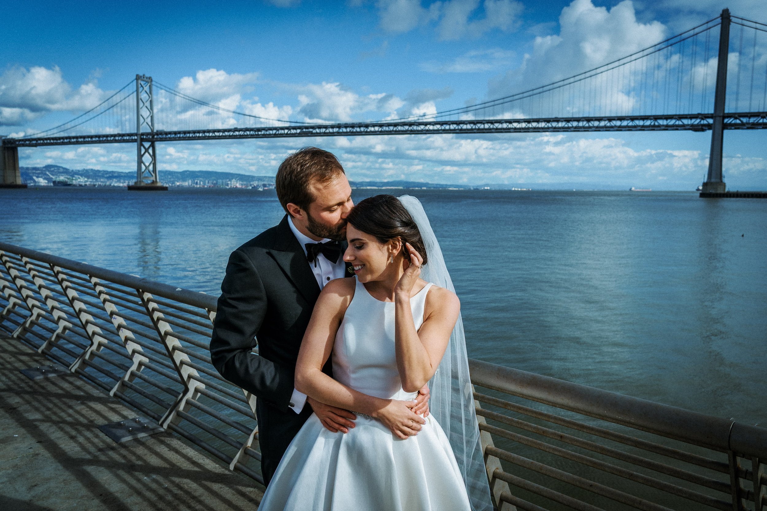 A newlywed couple embracing by the bay with the San Francisco Bay Bridge in the background.