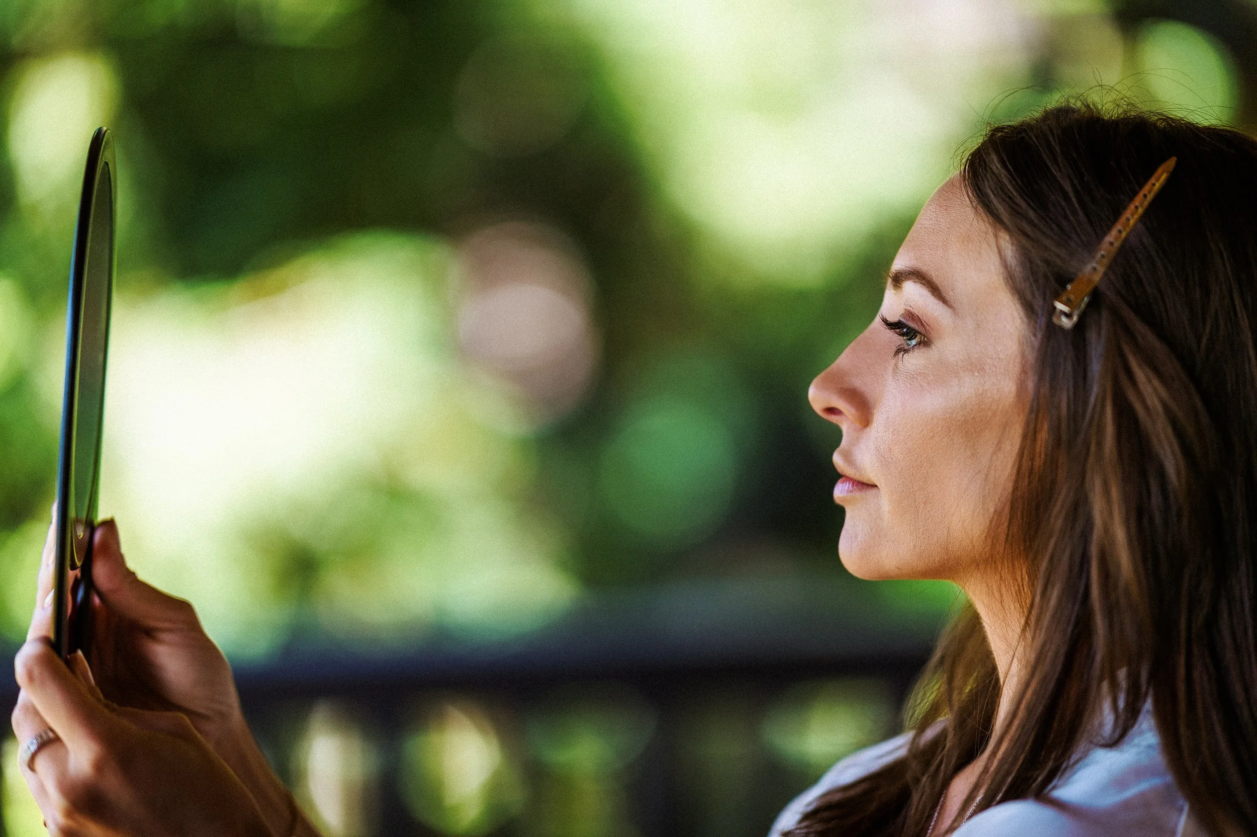 A bride with long brown hair, wearing a hair clip, is looking intently at her tablet, which she holds in her hands. The background is blurred with green and light bokeh effects.