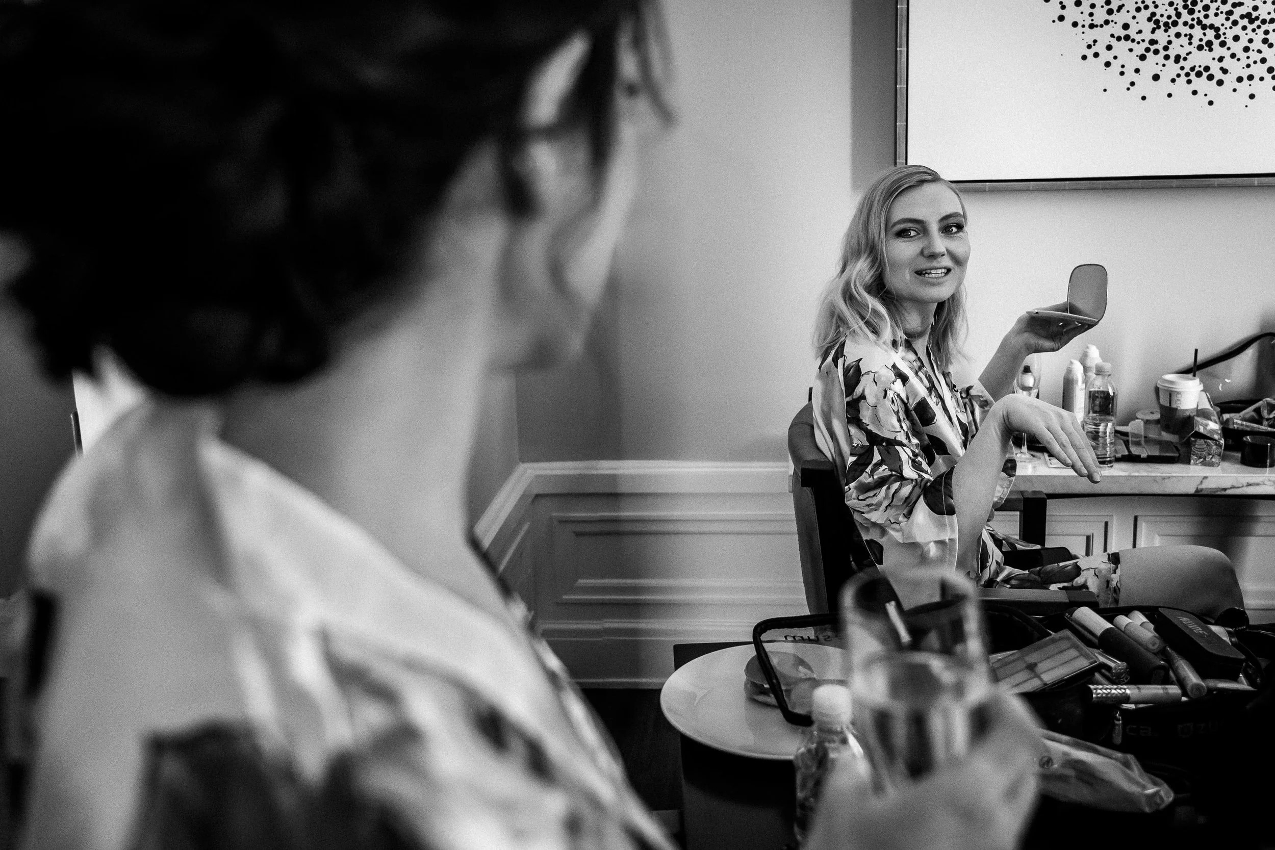 Black and white photo of two women in a room with makeup and beauty products. One woman in the foreground is slightly blurred, while the other woman in the background is sitting, holding a mirror, and smiling.