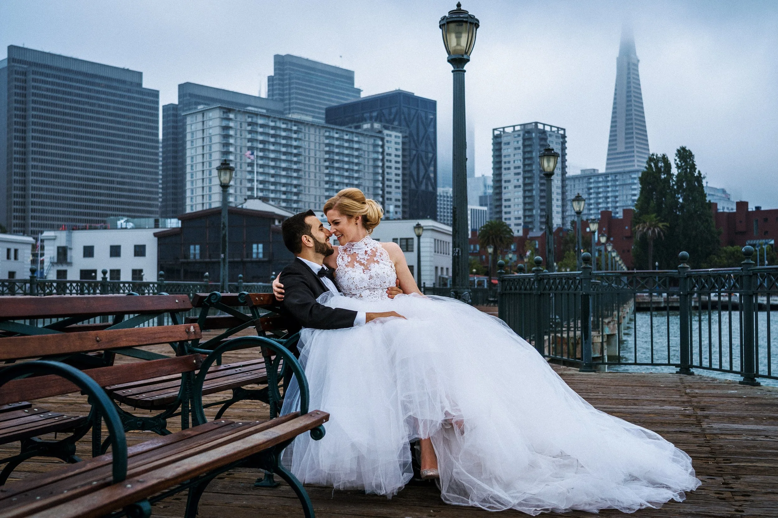 A couple in wedding attire sitting on a park bench on a pier, with San Francisco skyline and tall buildings in the background, embracing and smiling at each other.