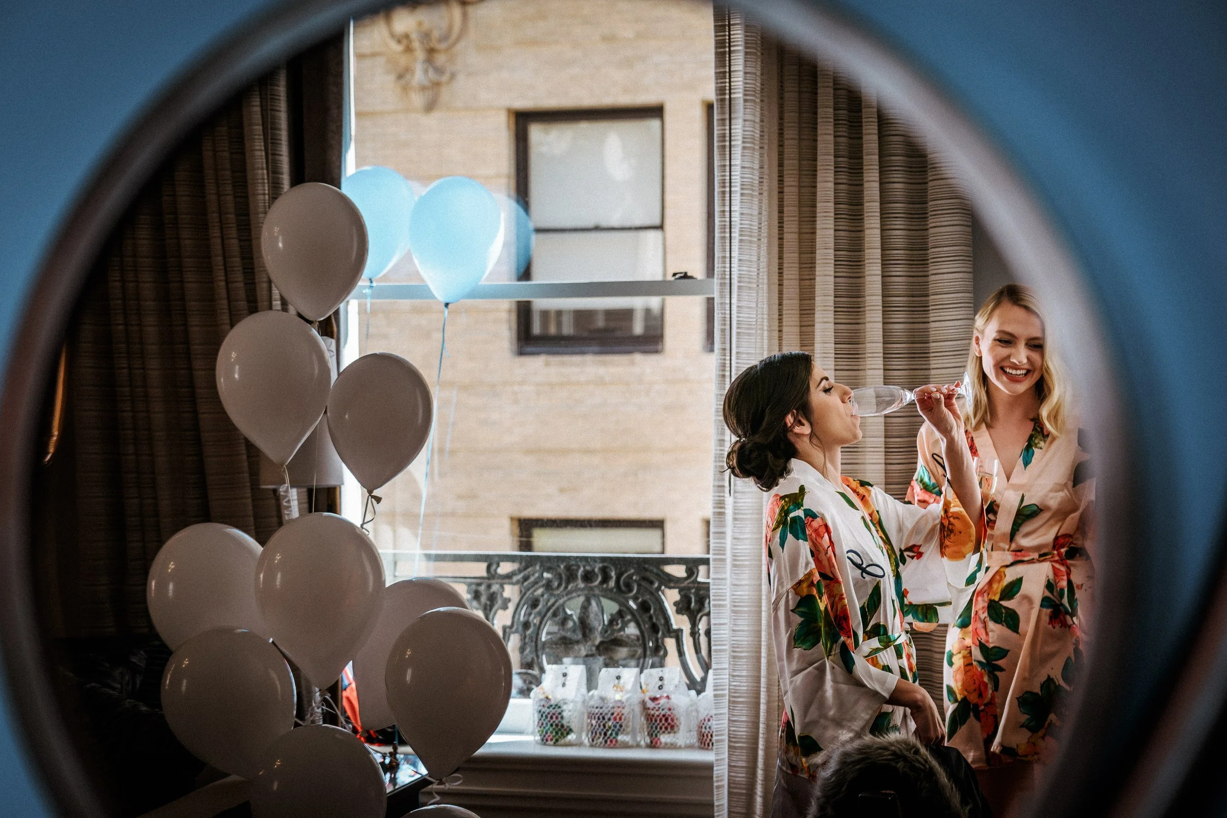 Women in floral pajamas celebrating indoors with balloons, one sipping from a champagne flute, seen through a laundry machine door
