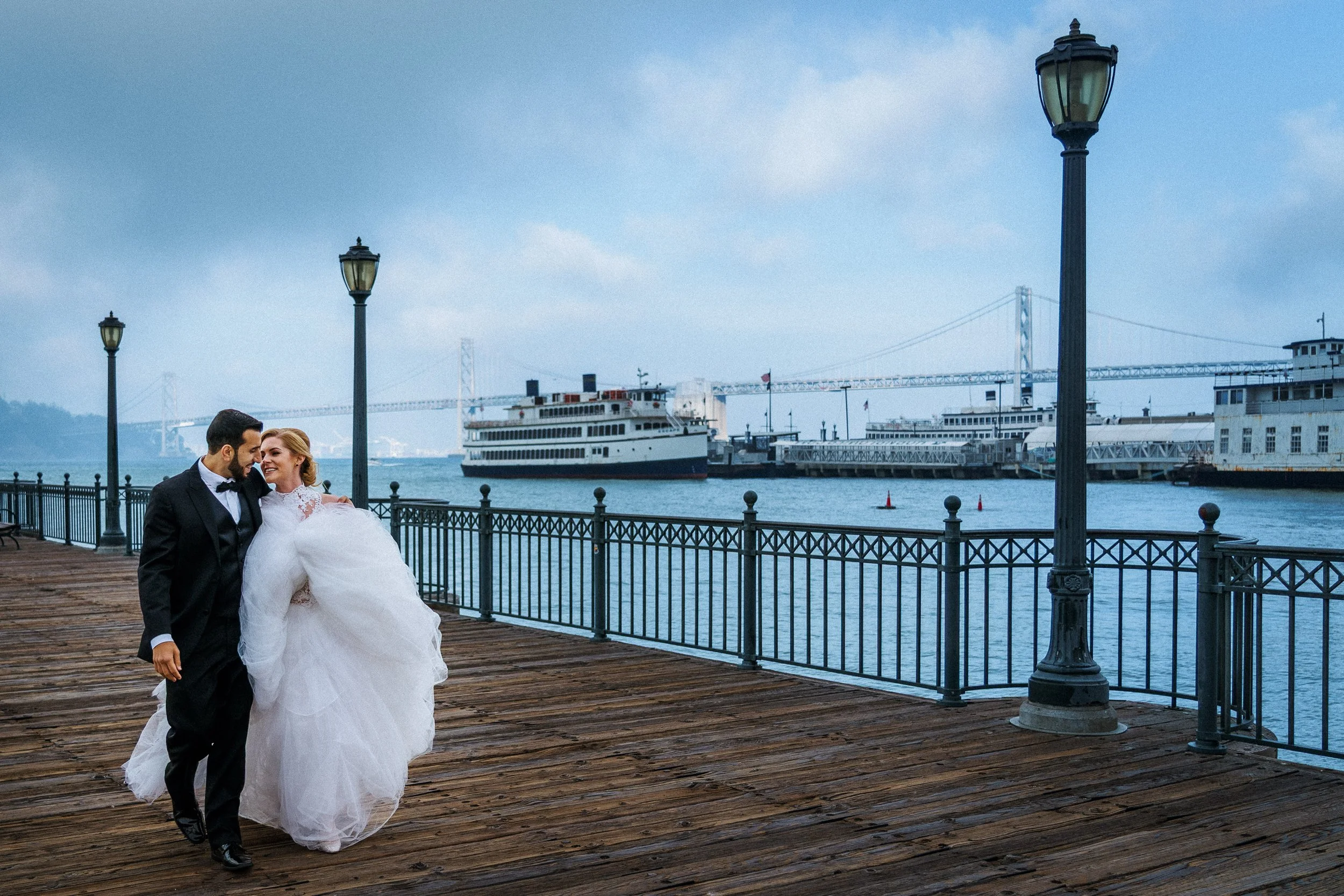 A newlywed couple dressed in wedding attire walking on a wooden pier with a scenic harbor view and a large ship in the background.