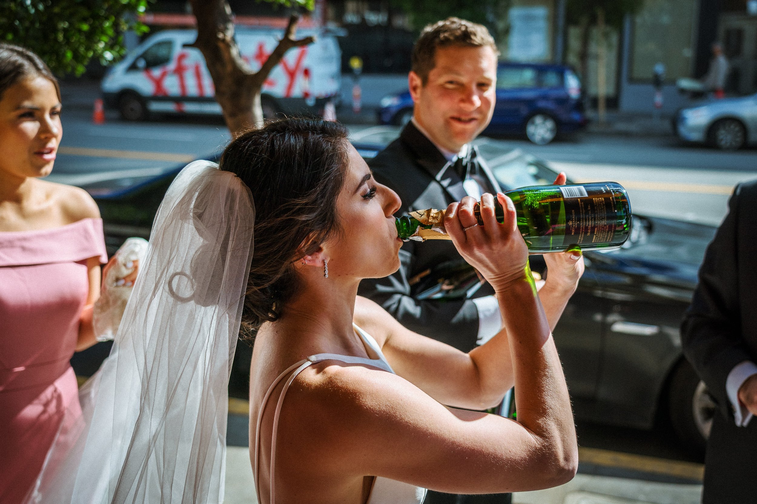 A bride in a wedding dress and veil drinks champagne from a bottle with a gold foil neck beside a groom in a black tuxedo, with a woman in a pink dress and another person in a black suit nearby, outdoors during daytime.