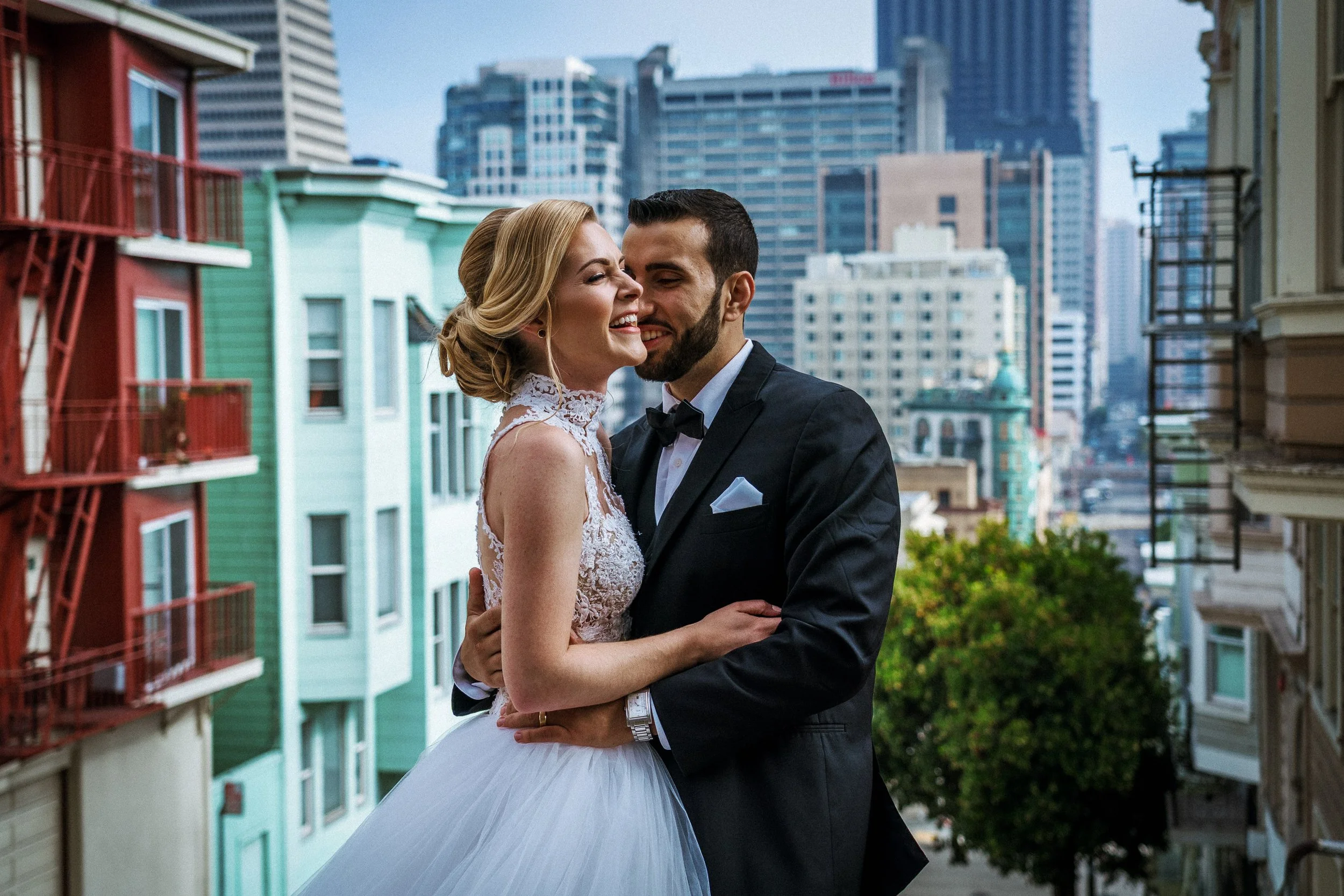 A couple dressed in wedding attire embracing on an outdoor balcony with a cityscape background of tall buildings and trees in San Francisco.
