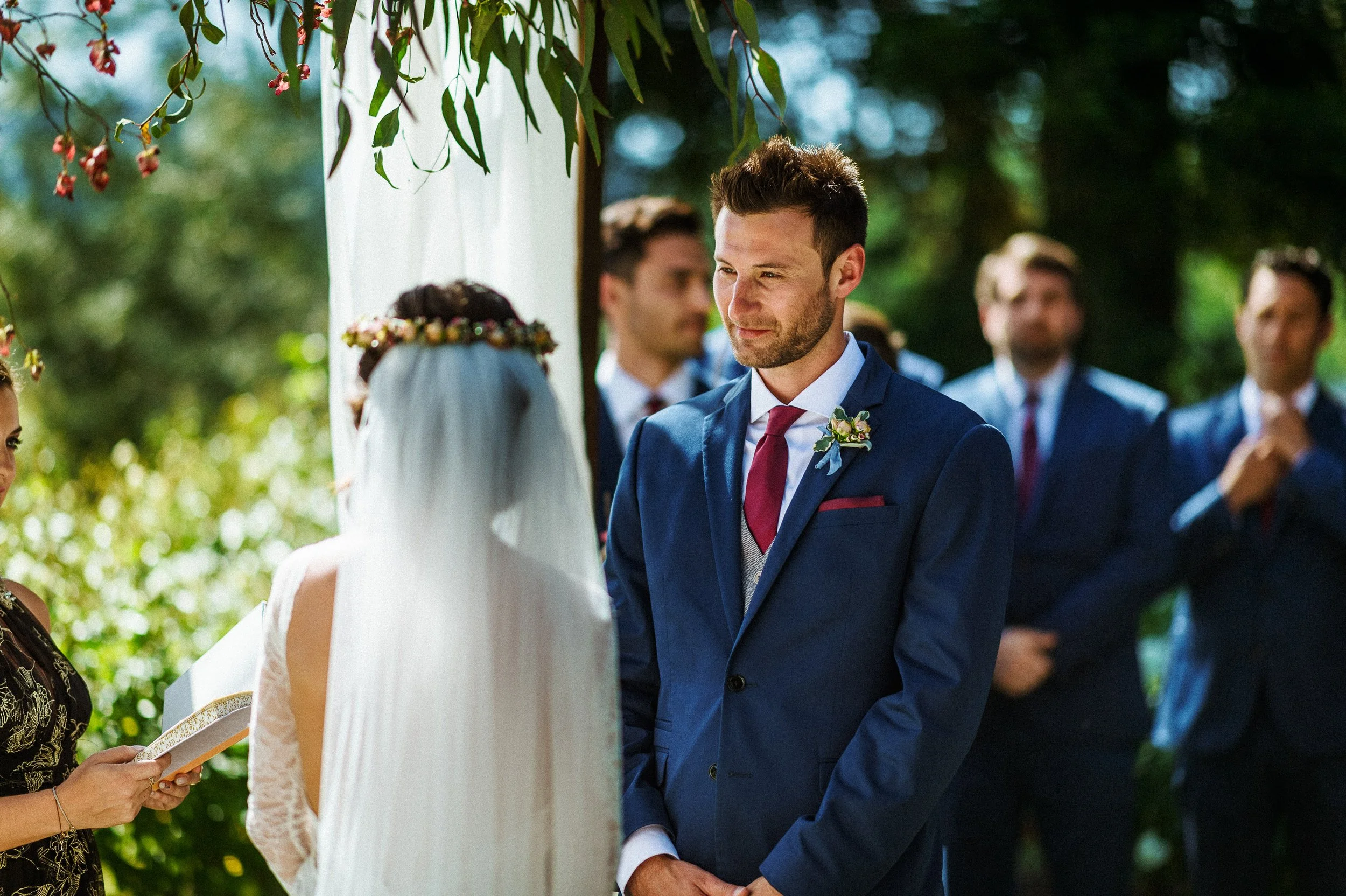 Groom and bride exchanging vows at an outdoor wedding ceremony, with bridal party in the background.