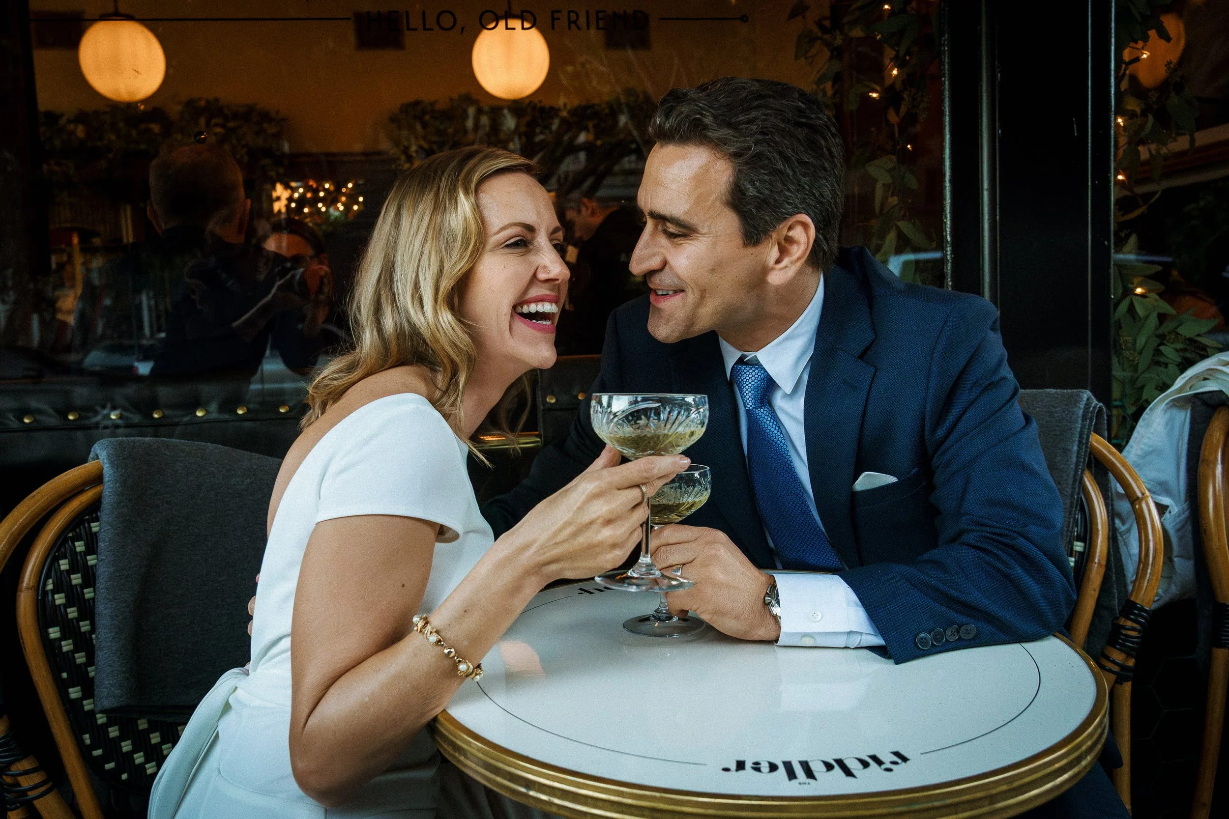 A joyful woman and man sharing drinks and laughing at a restaurant table. The woman wears a white dress and a bracelet, and the man is in a blue suit. They are enjoying each other's company.
