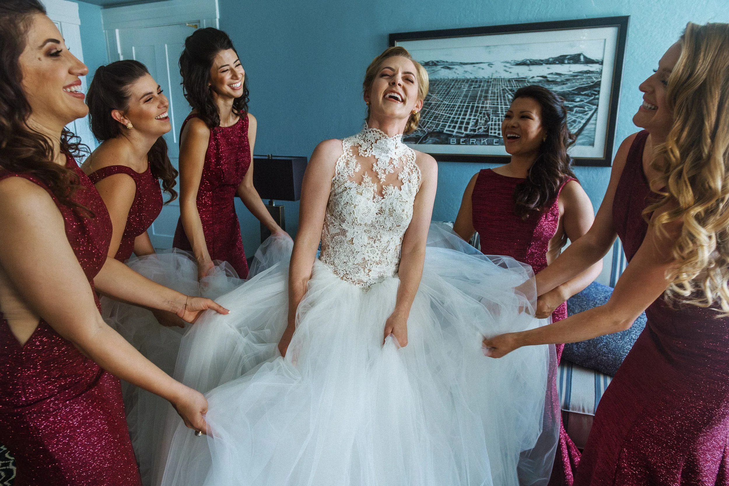Bride in a white lace wedding dress laughing with her eyes closed as her bridesmaids in matching burgundy dresses hold out her wedding gown in a room with teal walls and a landscape artwork.