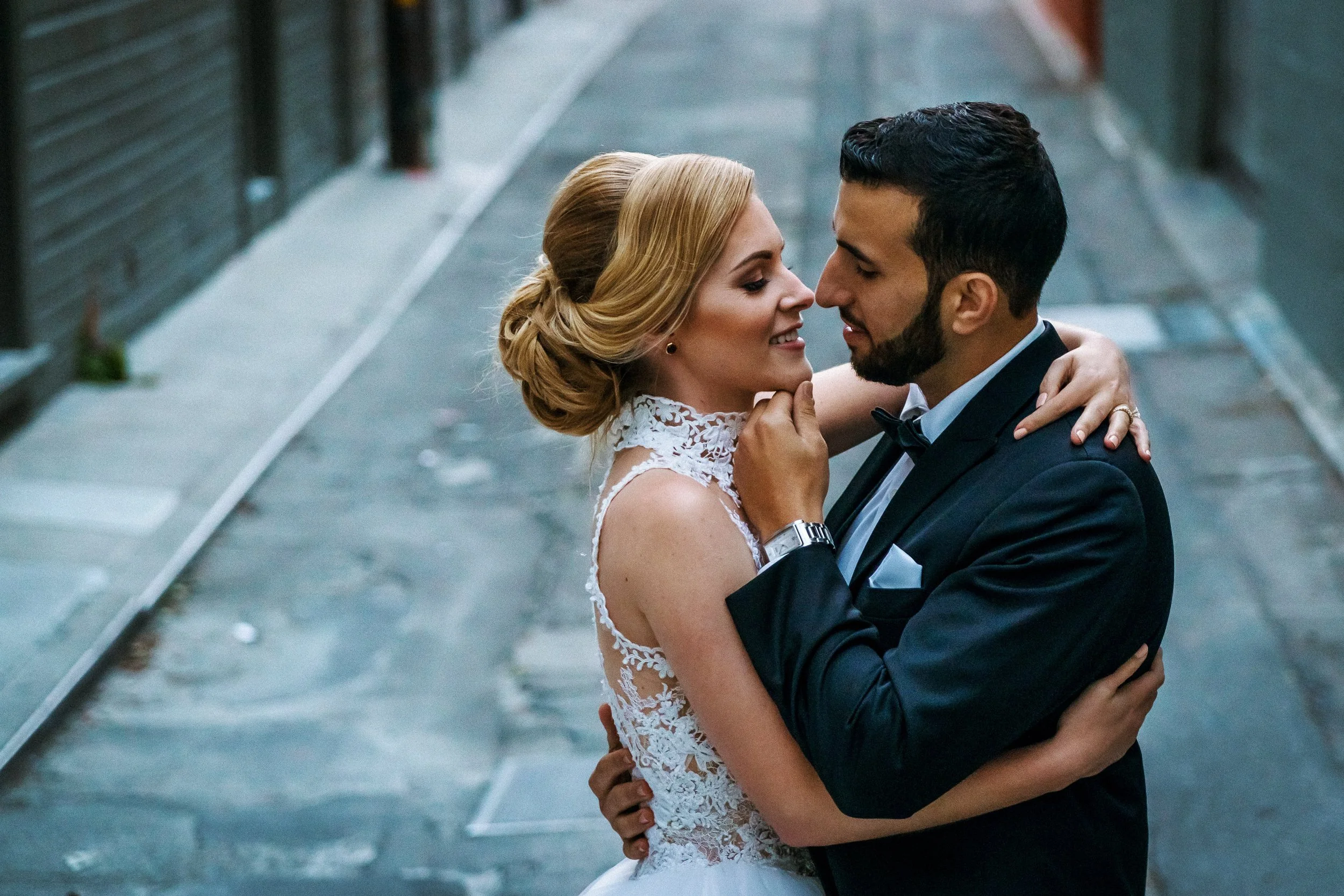 A bride and groom embracing close-up in a narrow outdoor alleyway, the bride with blonde hair in an updo wearing a lace wedding dress and the groom with dark hair and beard in a tuxedo, smiling gently at each other.