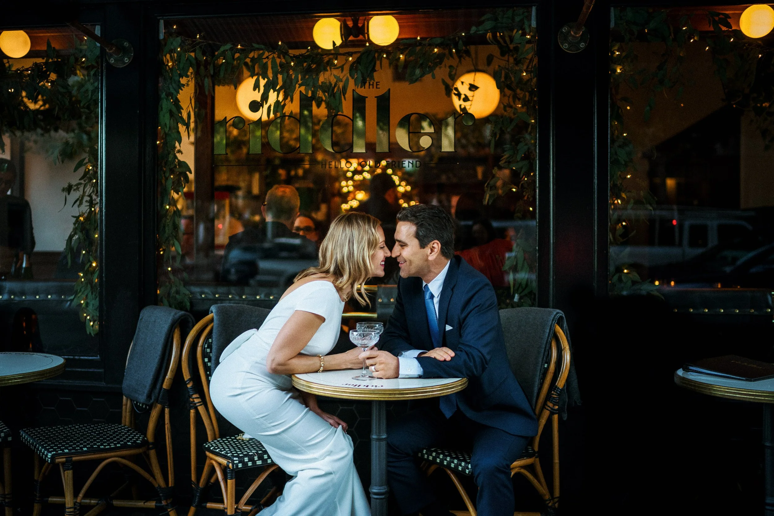 Couple having an intimate moment at a small round table inside a dimly lit restaurant. The woman in a white dress and the man in a dark suit lean close, smiling and holding a champagne glass. The window behind them displays the restaurant's name 'The