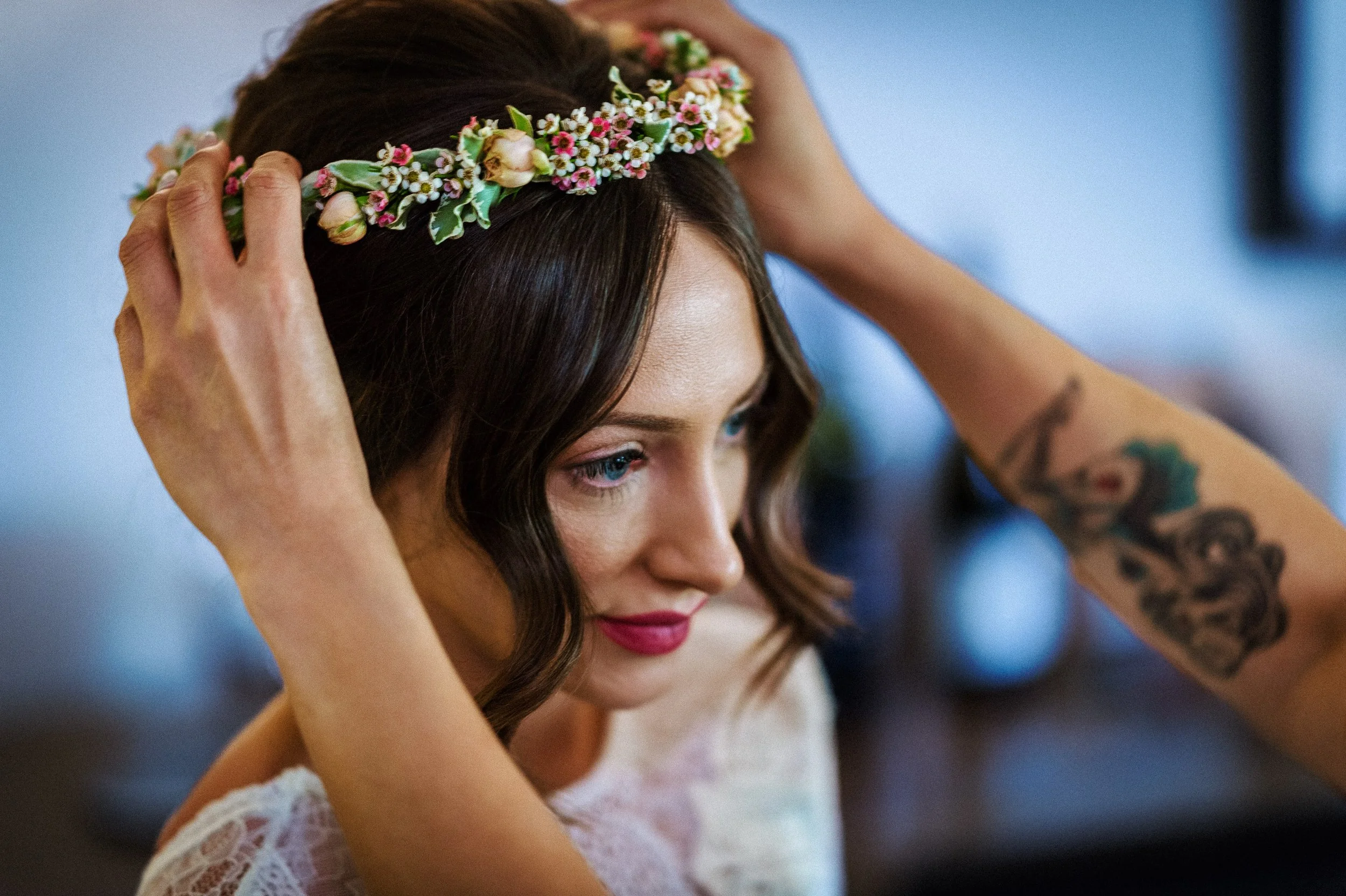 A bride with brunette hair and pink lipstick, wearing a flower crown and a lace top, adjusting her flower crown with her hands.