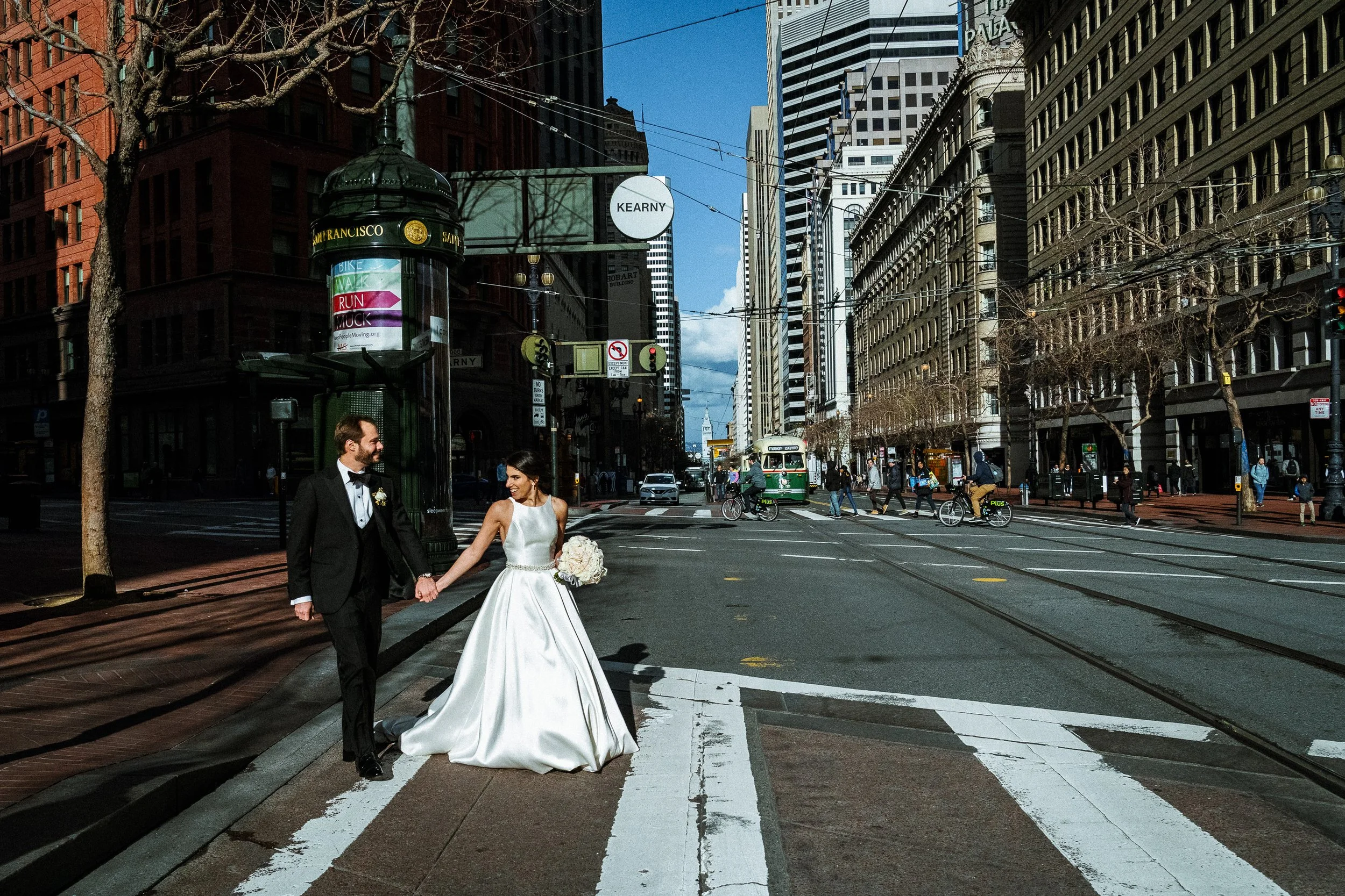 A bride and groom holding hands on a city crosswalk, with tall buildings and a streetcar in the background.