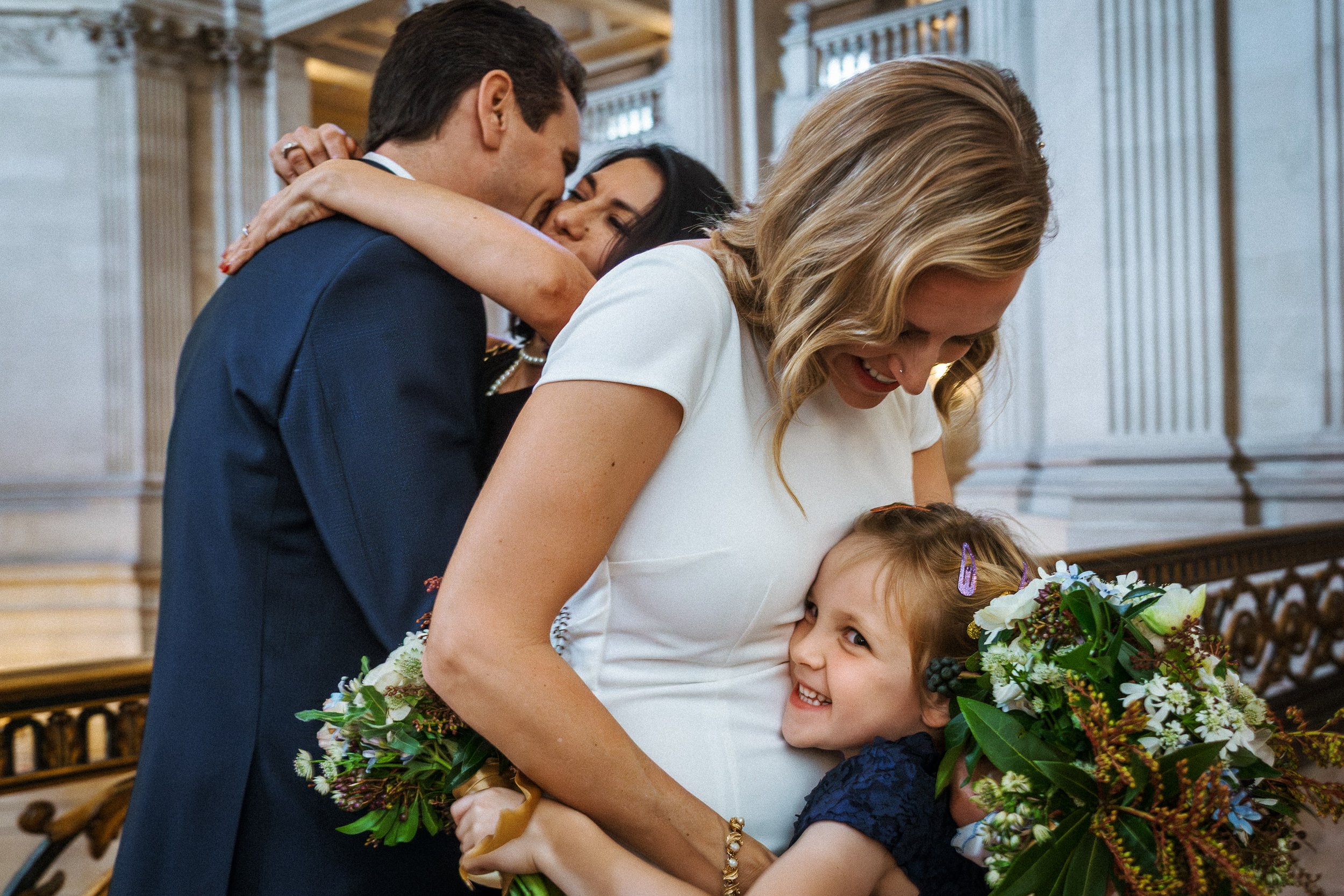 A woman with blonde hair in a white dress hugging a young girl with light brown hair and a purple hair clip, both smiling, with the woman holding a bouquet of flowers. Behind them, a man in a navy suit hugging a woman with black hair.