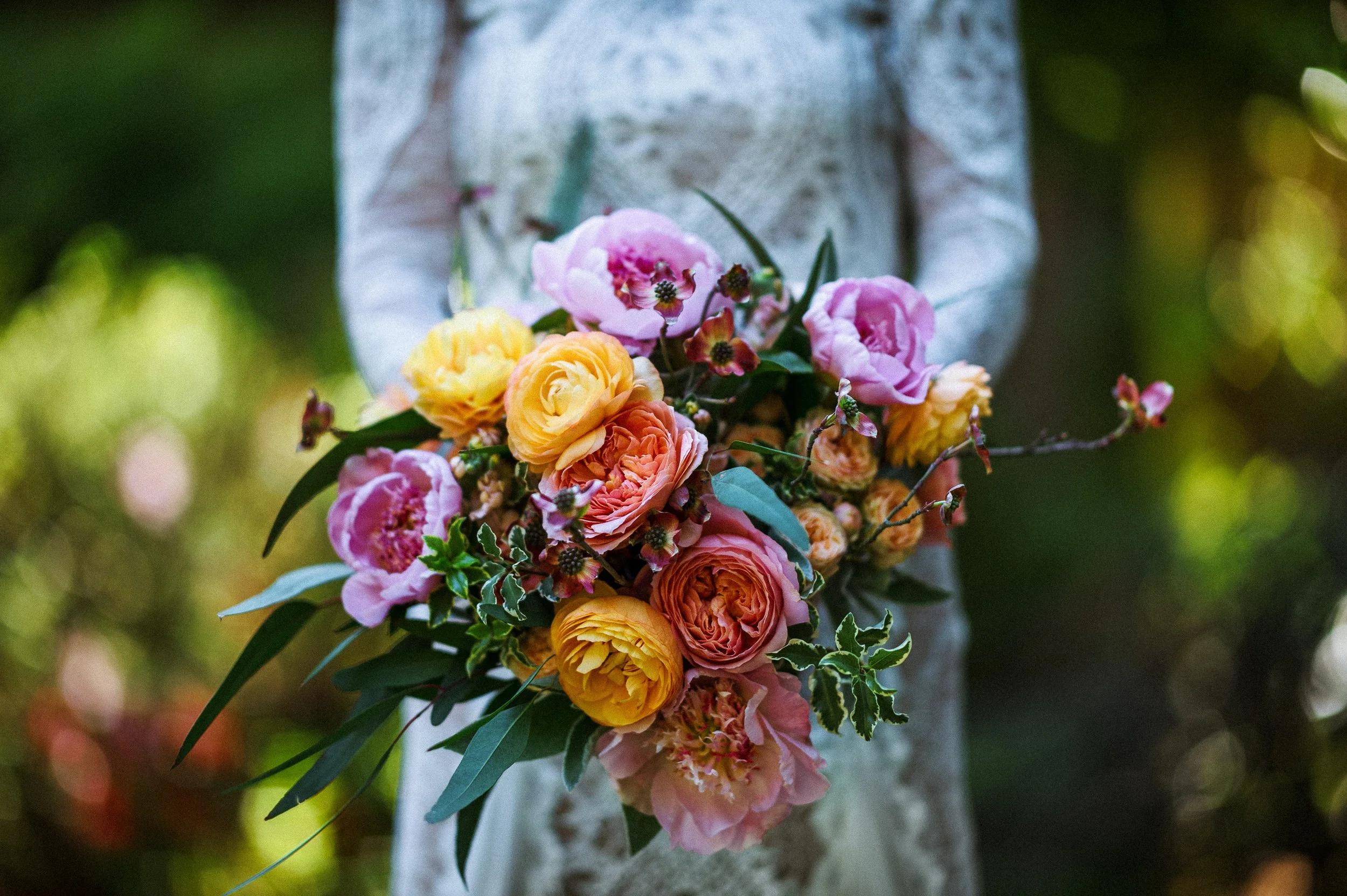 A woman in a white lace dress holding a colorful bouquet of pink, yellow, and orange flowers.