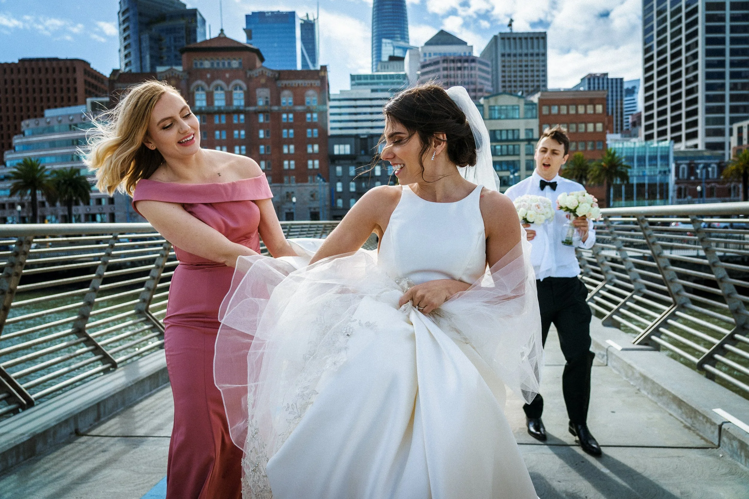 A bride in a white wedding dress, smiling, is being assisted by a bridesmaid in a pink dress. A groomsman holding a bouquet is in the background on a city bridge with a skyline of tall buildings.