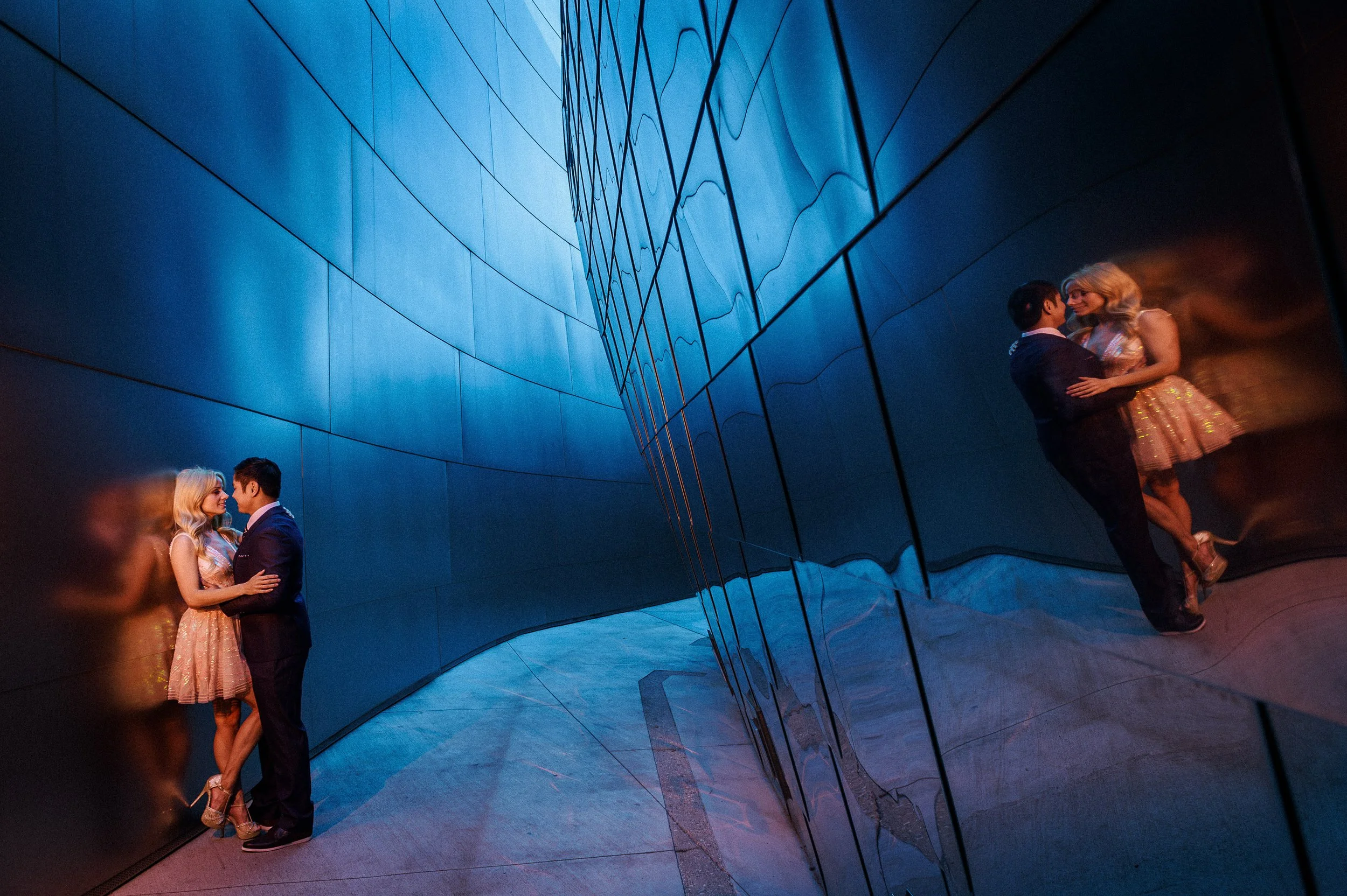 A couple sharing a romantic moment with their foreheads touching at the Walt Disney Concert Hall in Los Angeles. Their reflections can be seen on the curved, shiny blue surface.