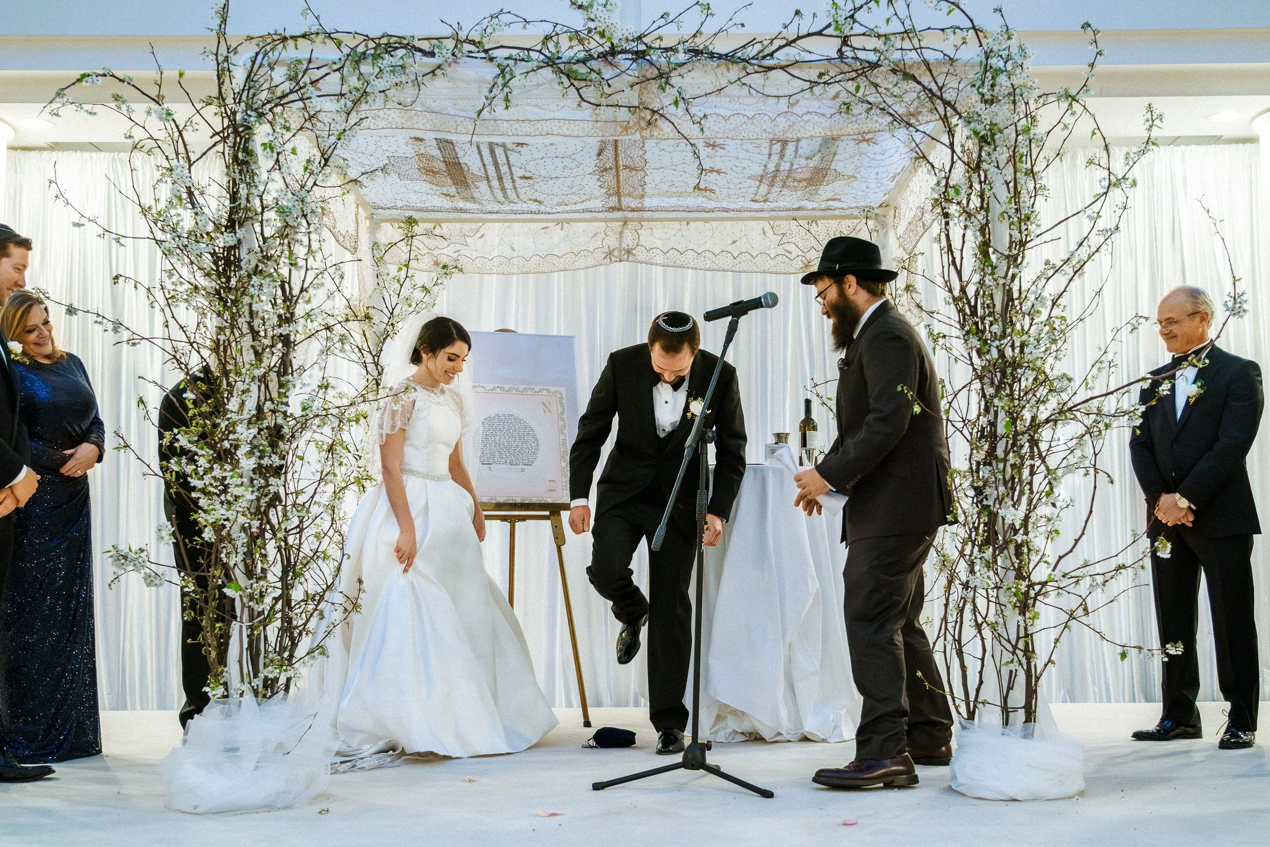 Jewish wedding ceremony with couple and guests standing under a decorated arch of bare branches with white flowers, officiant and microphone in the center, transparent curtains in the background.