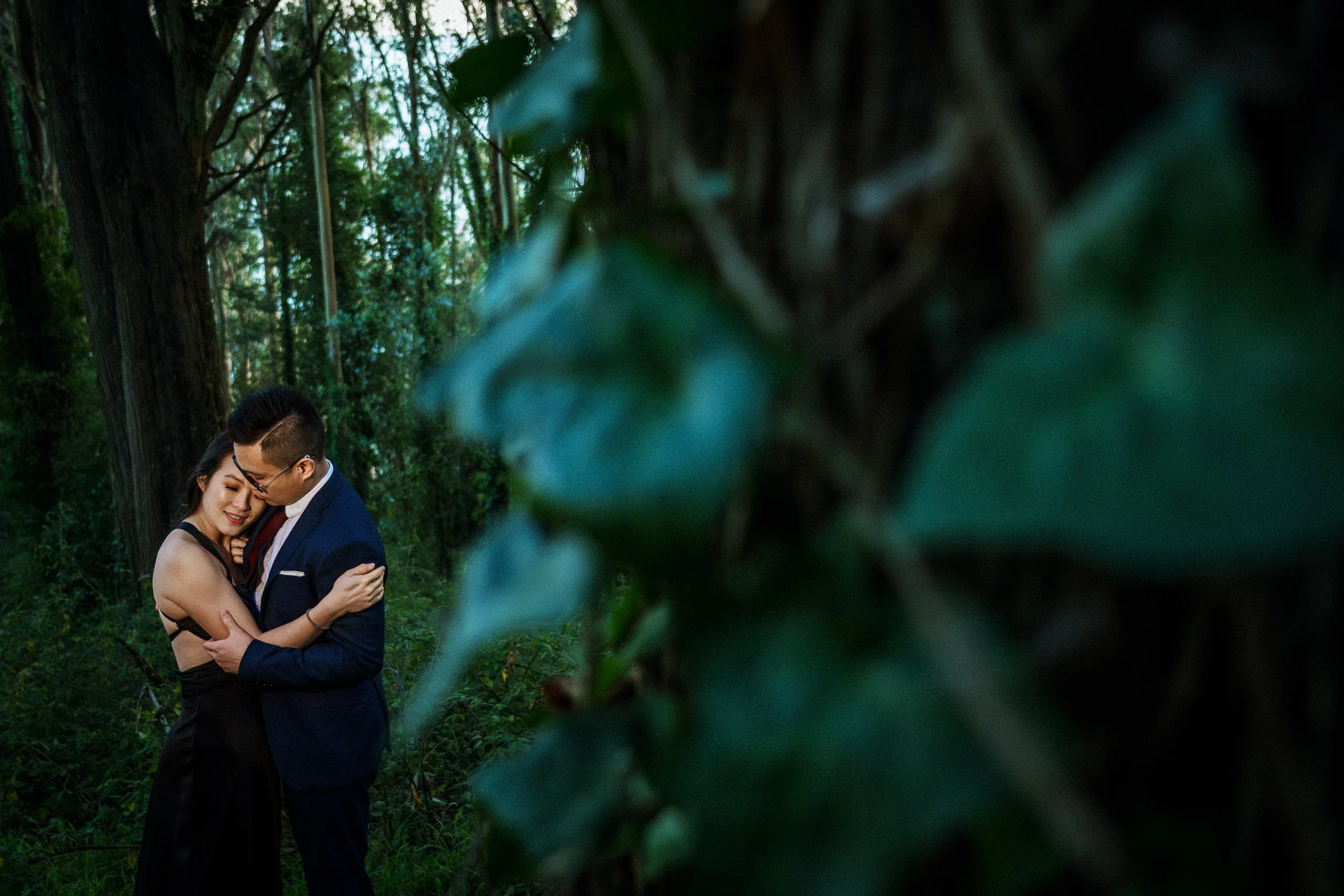 A romantic couple hugging in a forest, with large green leaves in the foreground and tall trees in the background.