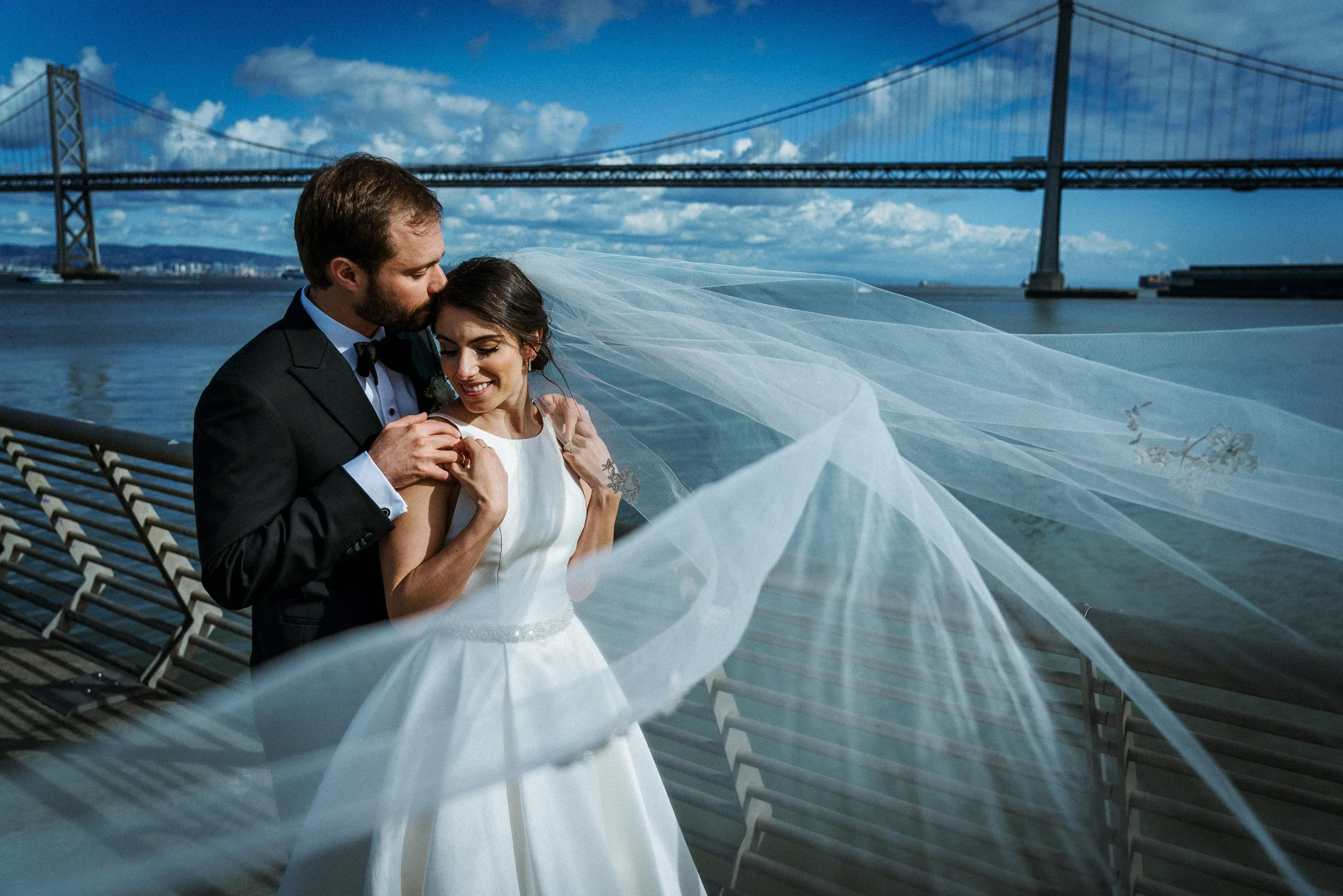 A newlywed couple standing by a waterfront with a bridge in the background. The groom is in a black tuxedo and the bride is in a white wedding dress with a long veil. The groom gently kisses the bride's forehead while she smiles, holding her hands ne
