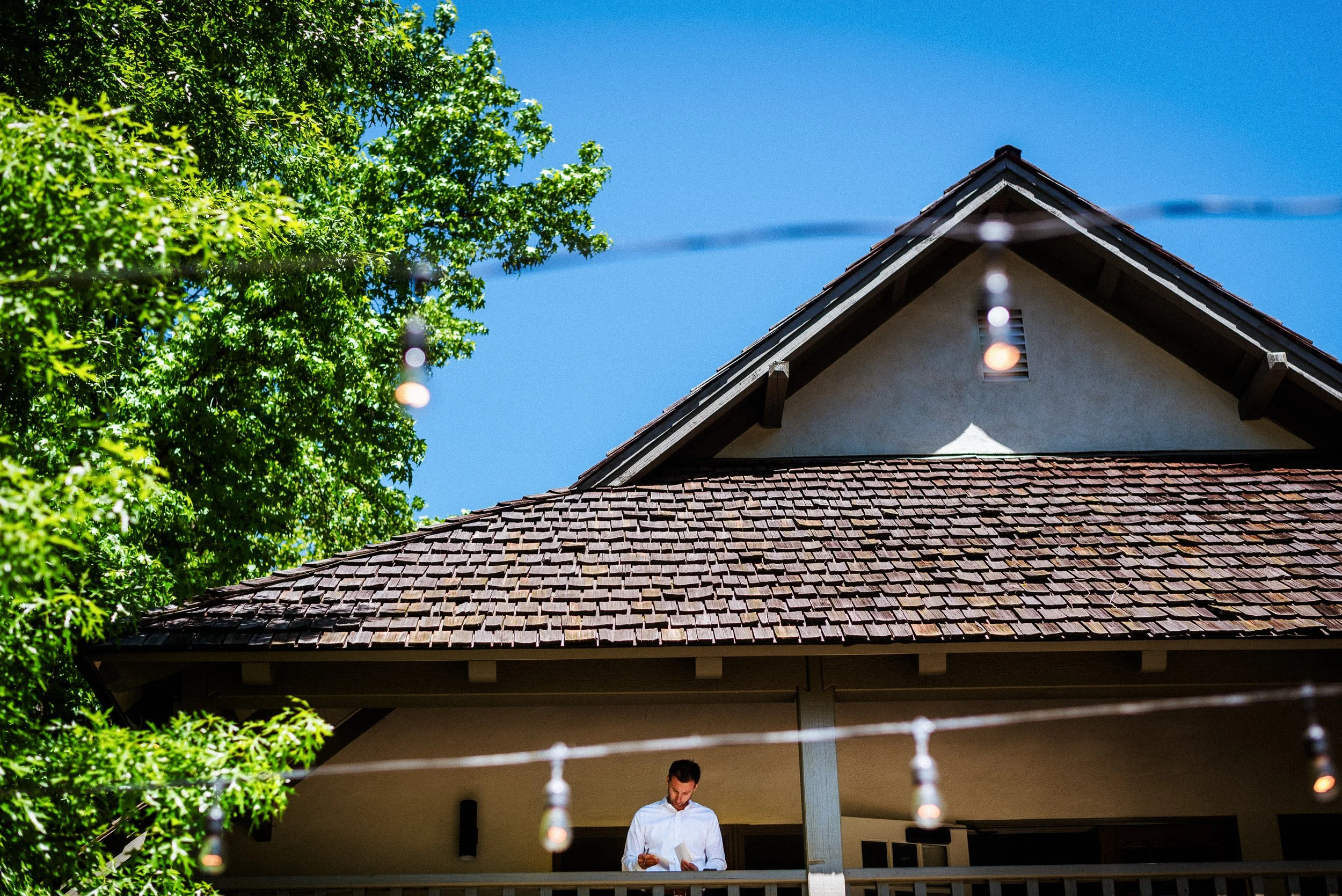 A groom standing on a balcony under a house with a brown tiled roof, green trees, and string lights, with a clear blue sky in the background.