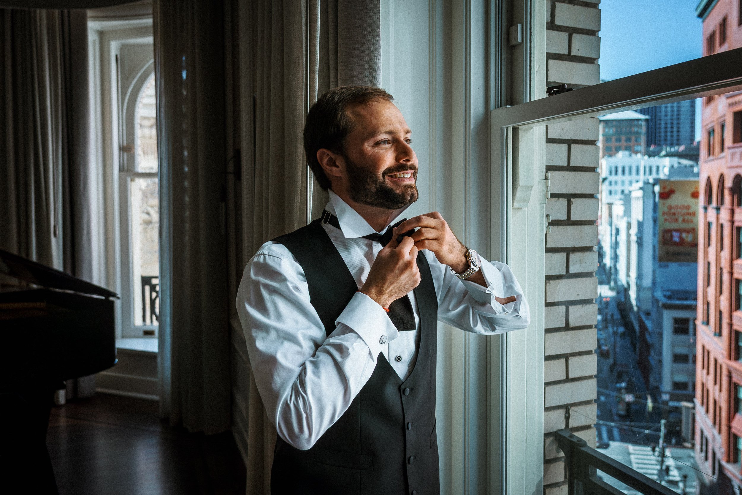 A groom in tuxedo adjusting his bow tie by a window in an urban apartment, getting ready for the wedding day.
